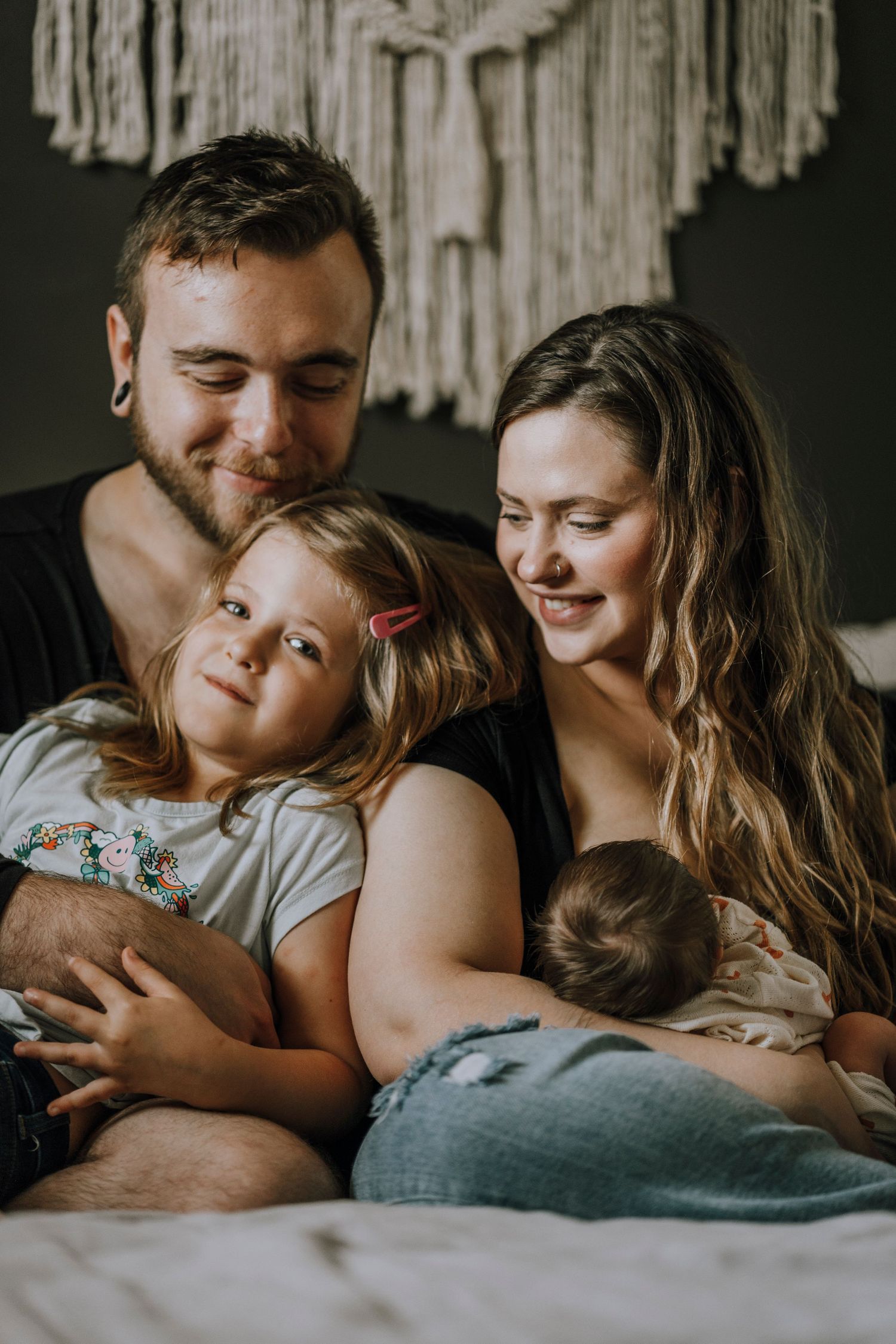 A young family shares an intimate moment together on their bed with a macrame backdrop.