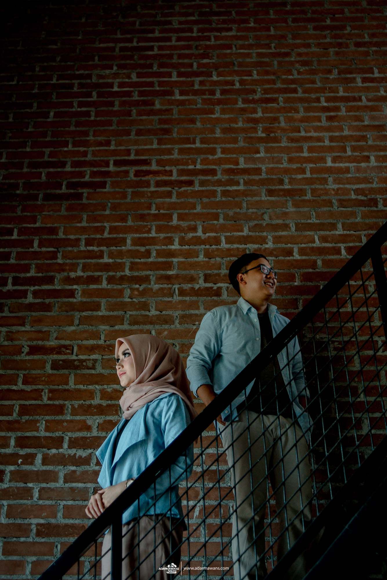A couple stands on an industrial staircase against a brick wall in atmospheric lighting.