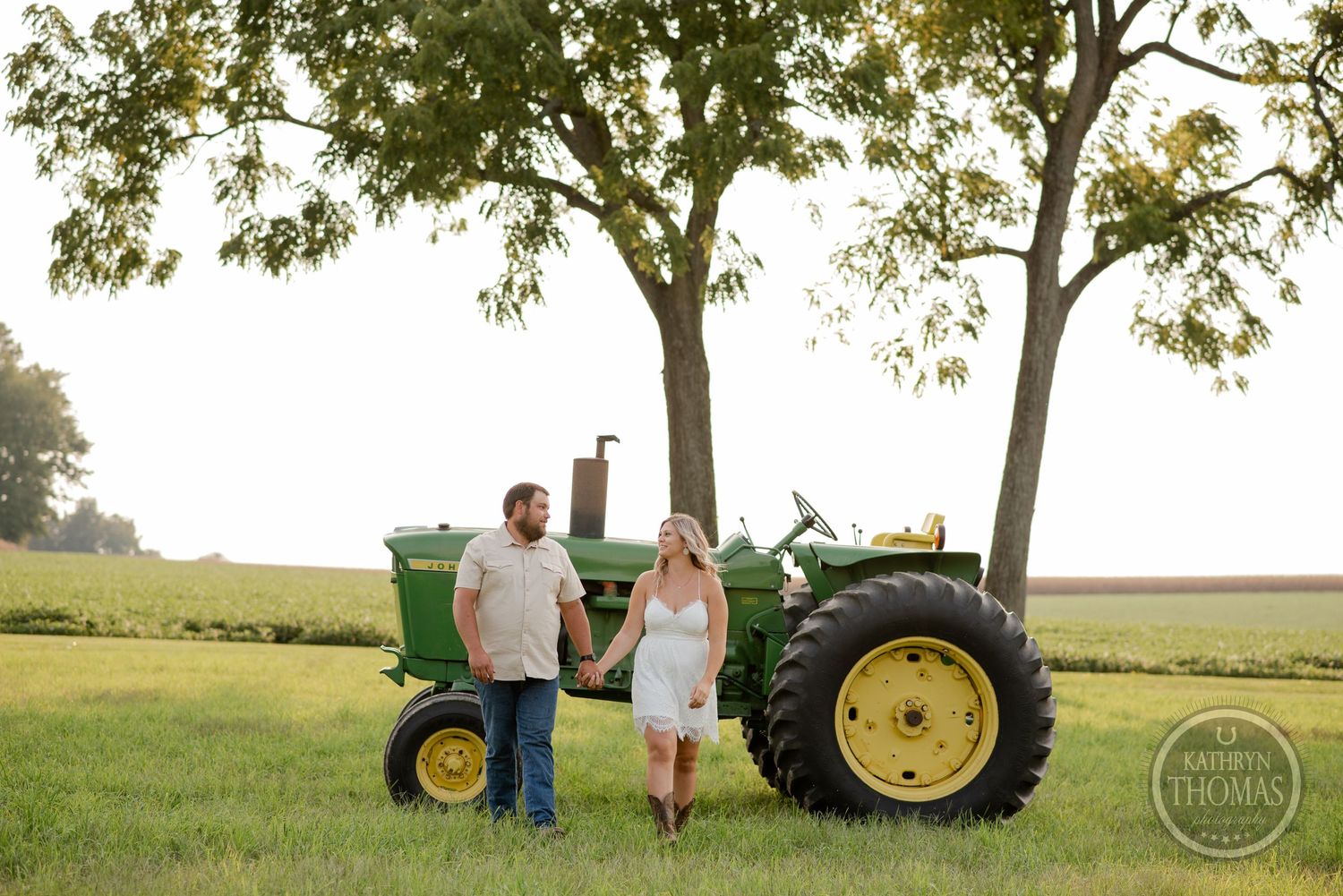 Couple stands together beside classic John Deere tractor with trees in background.