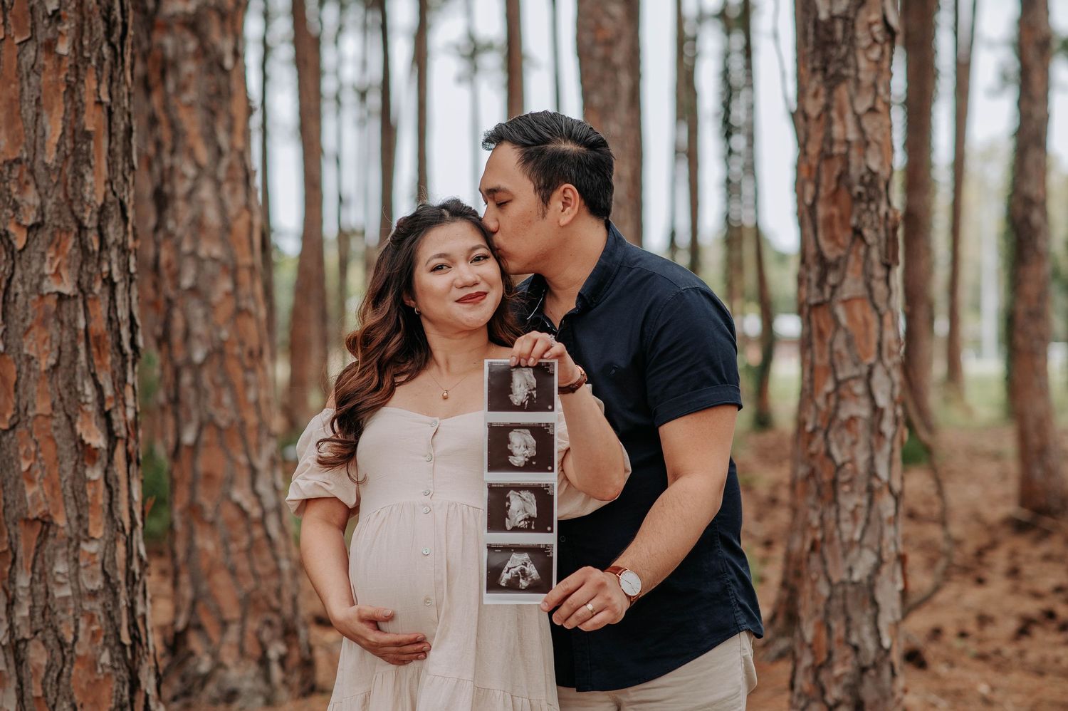 A couple poses in a pine forest while holding ultrasound photos.