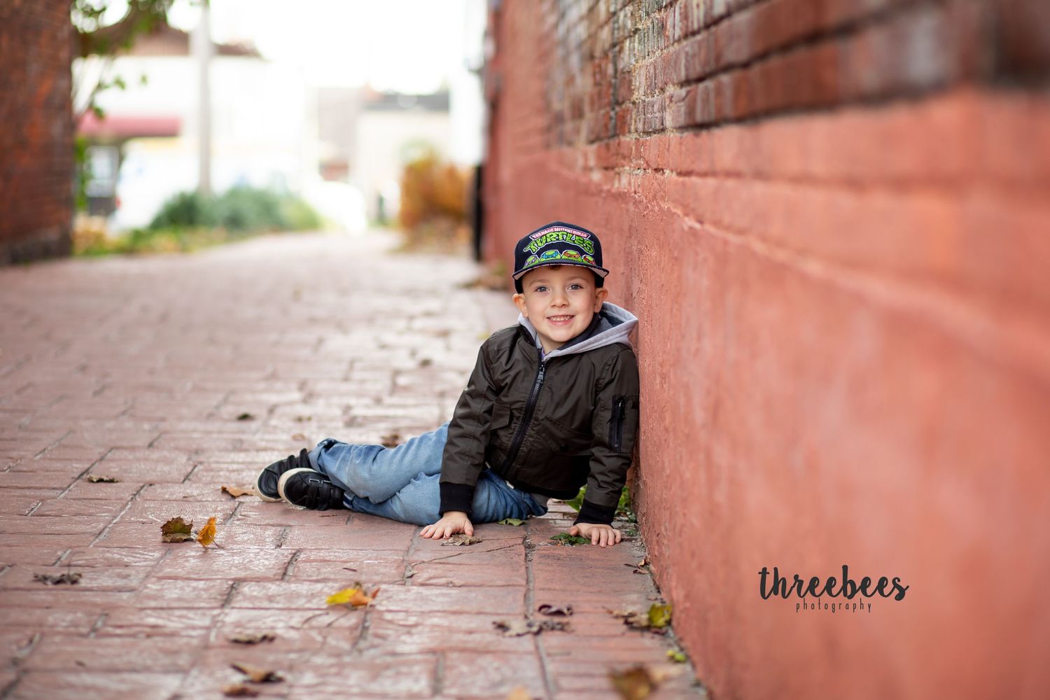 Child in baseball cap sitting against red brick wall with fallen autumn leaves.
