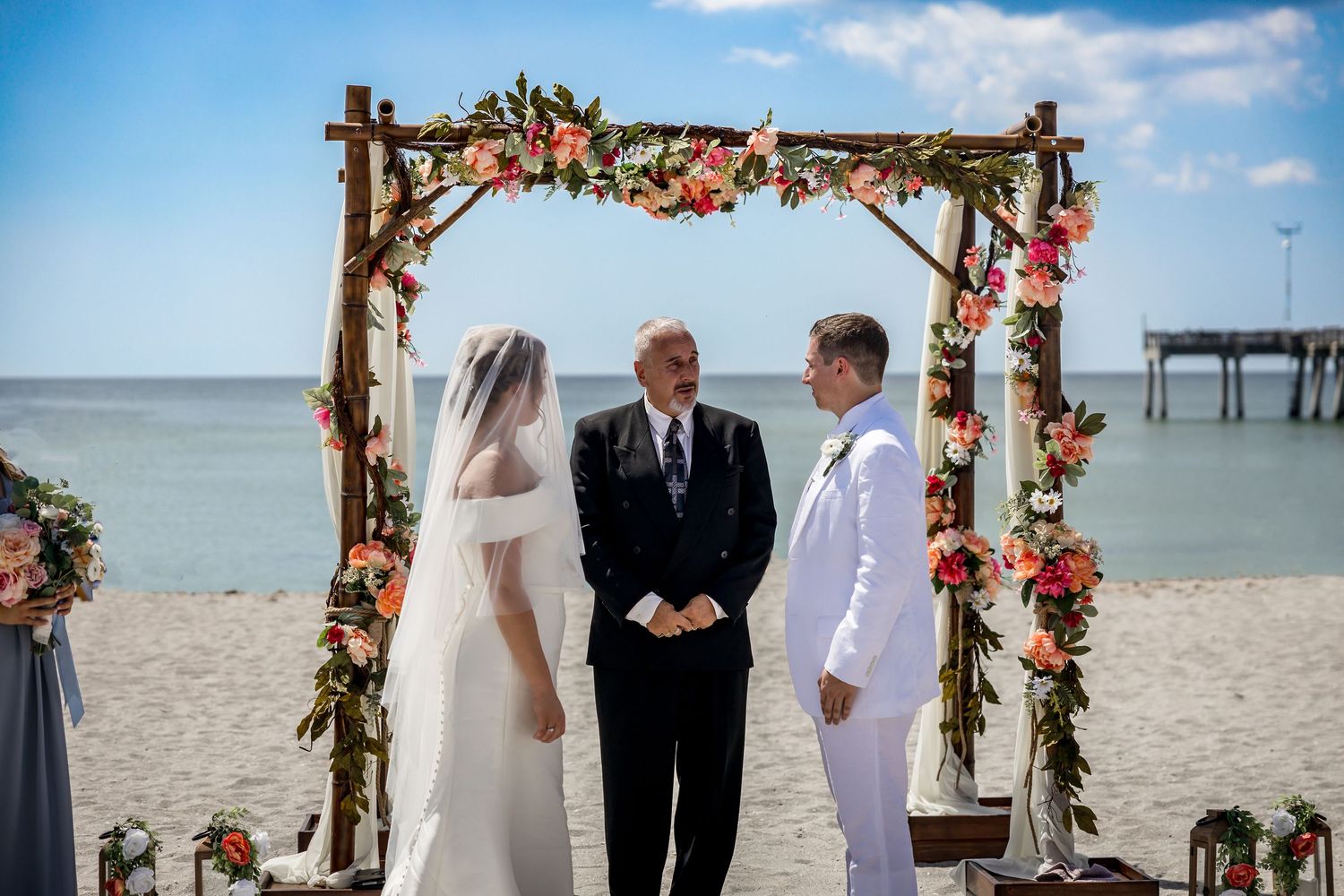 Bride and groom exchange vows at a beach ceremony under a rustic wooden arch decorated with pink and coral flowers.