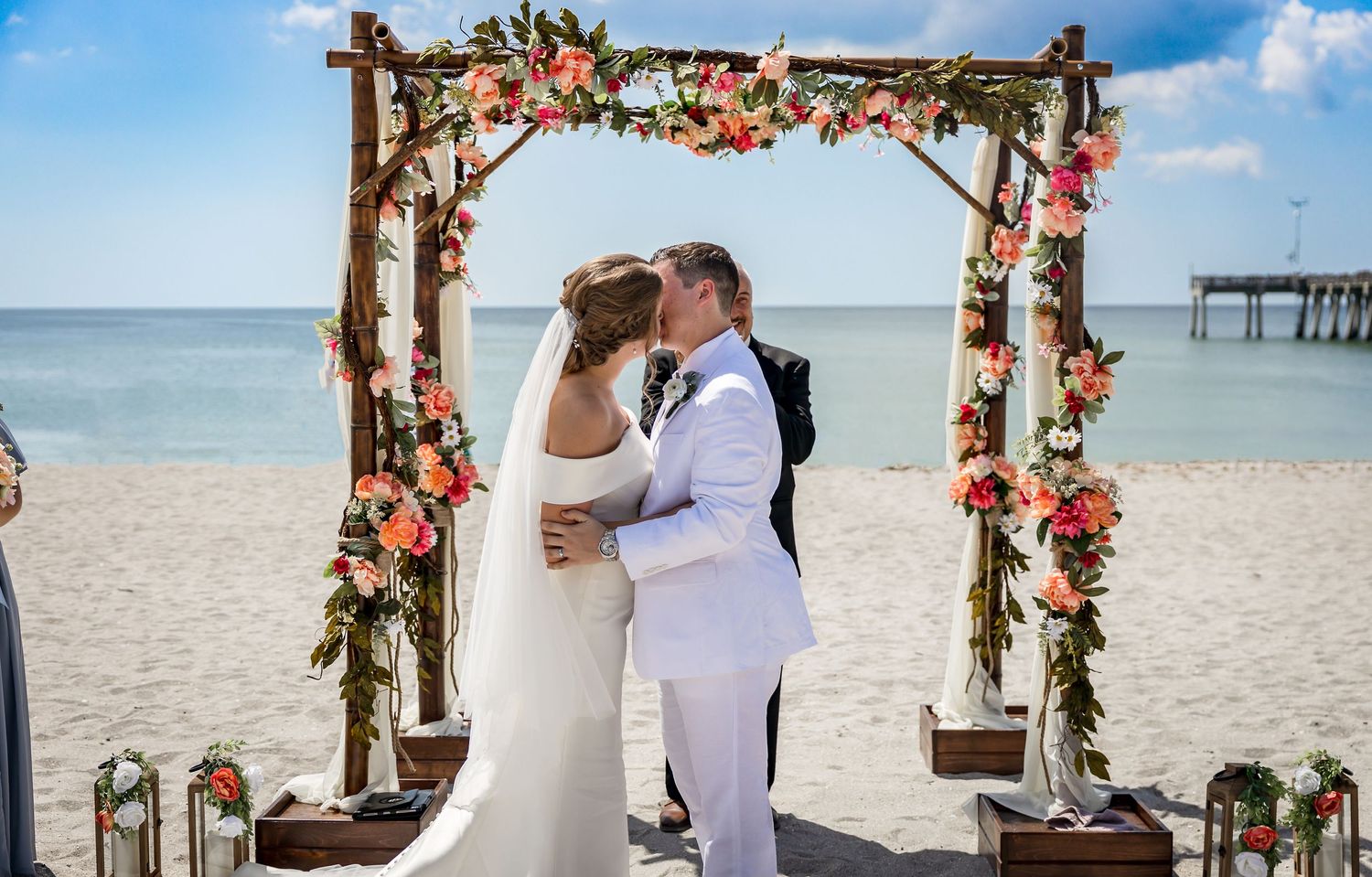Bride and groom share first kiss under floral arch at beachside wedding ceremony.