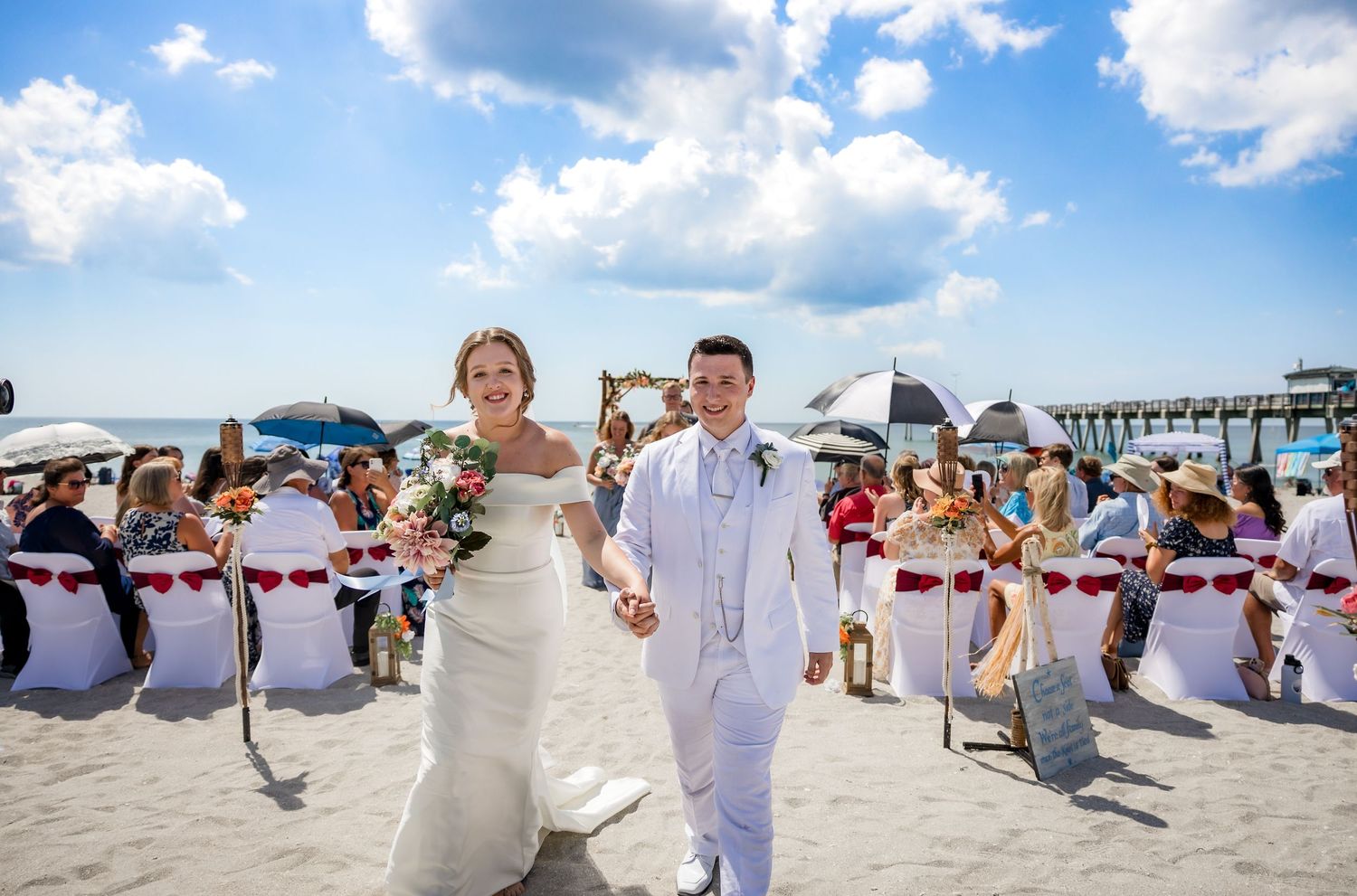 Newlyweds walk joyfully down beach aisle after outdoor seaside wedding ceremony.