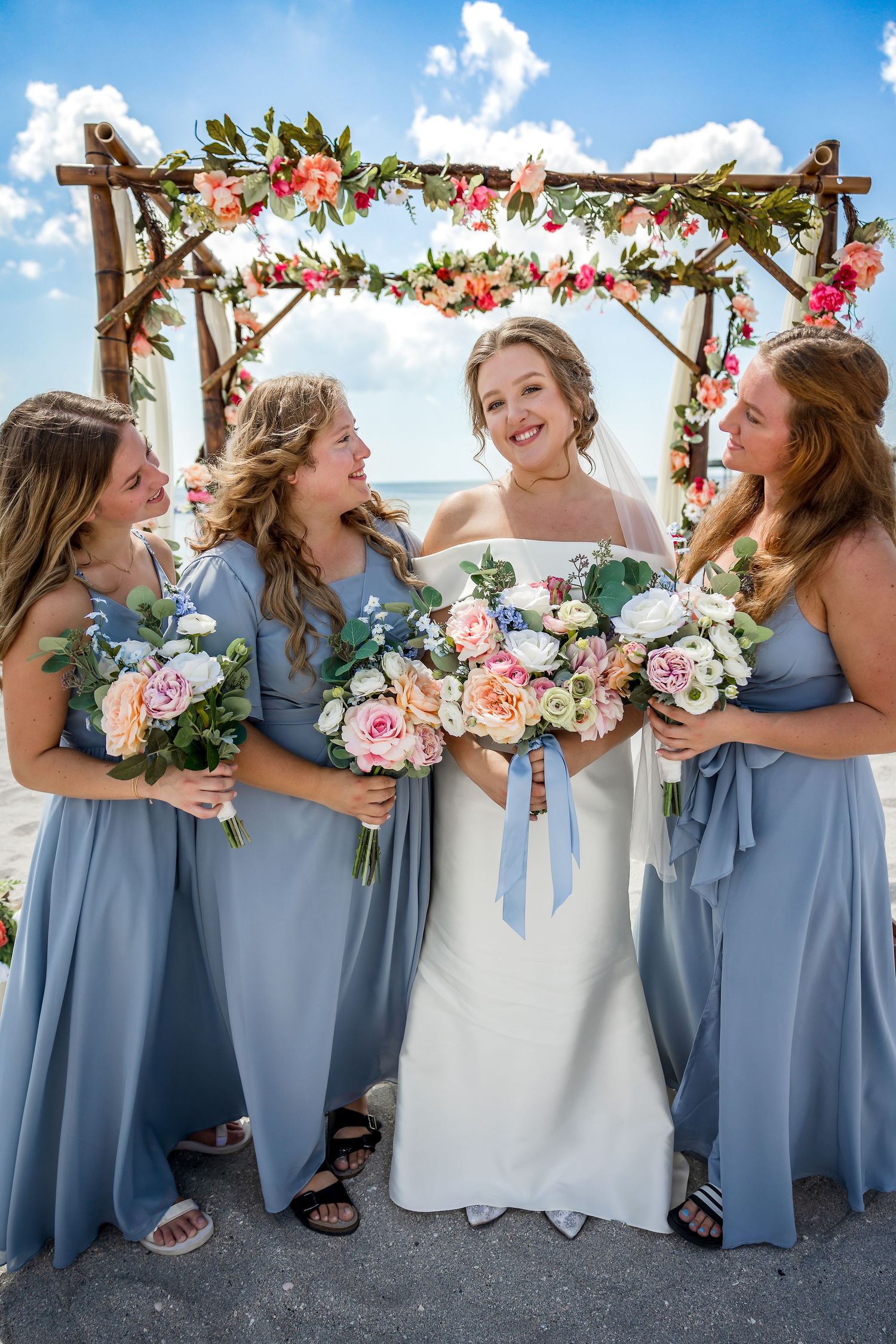 Bride smiles with three bridesmaids in dusty blue gowns holding floral bouquets at beach.