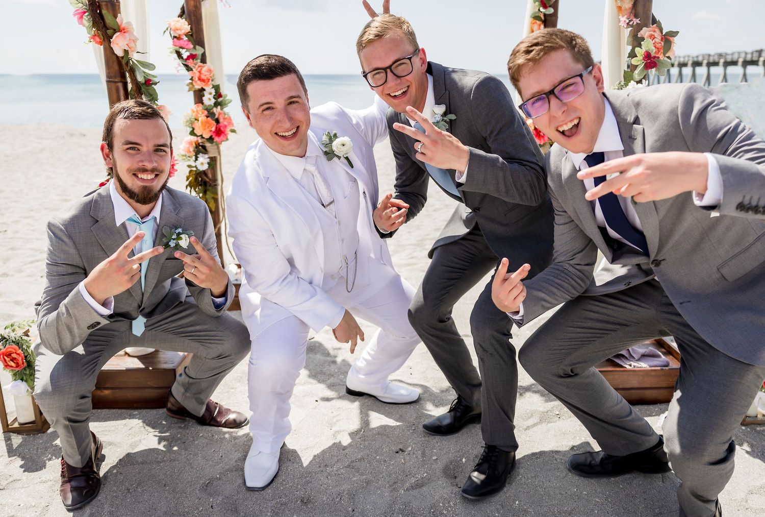 Groom in white suit poses playfully with groomsmen in gray suits at beach wedding.
