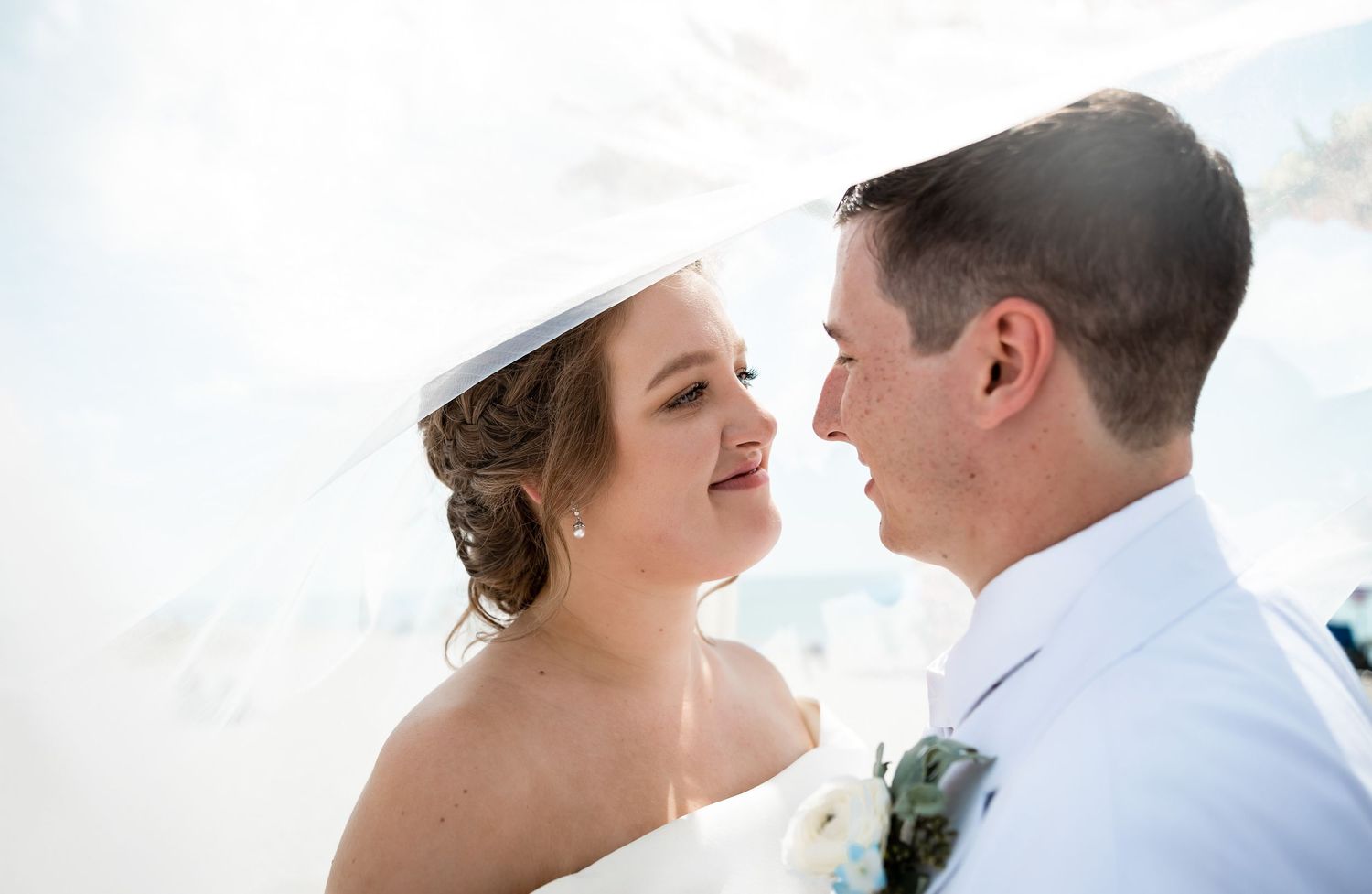Bride and groom share romantic moment under flowing veil on sunny beach.