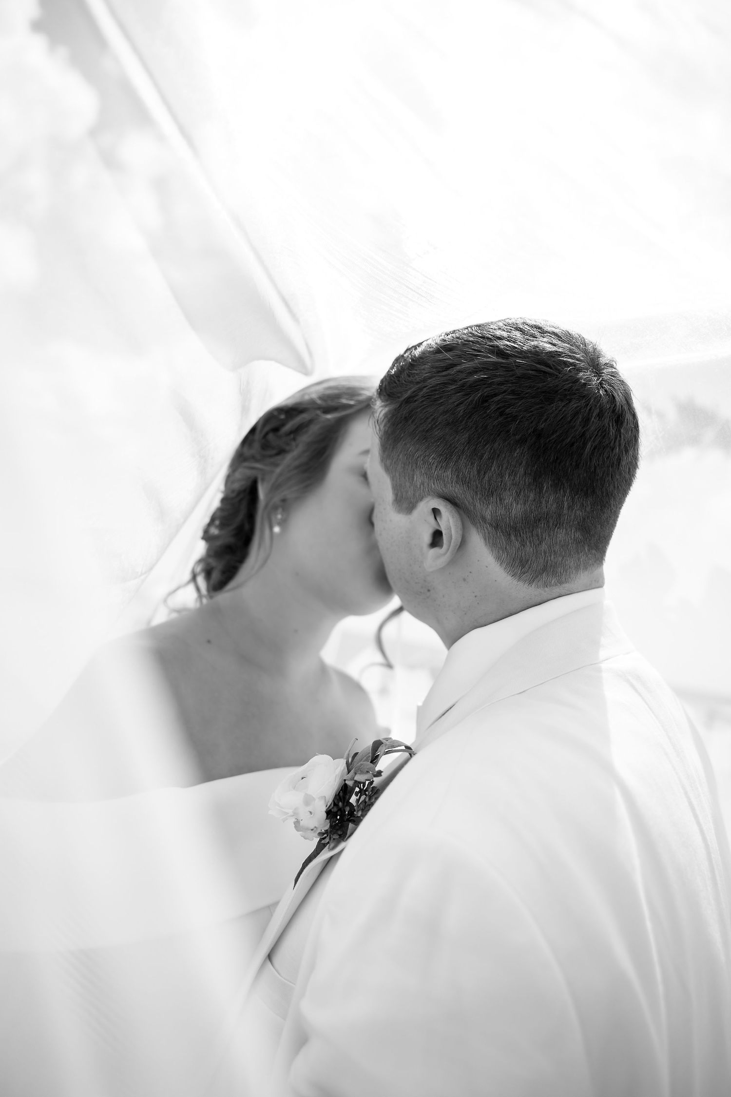 Black and white photo of a bride and groom sharing a romantic kiss beneath a flowing veil.