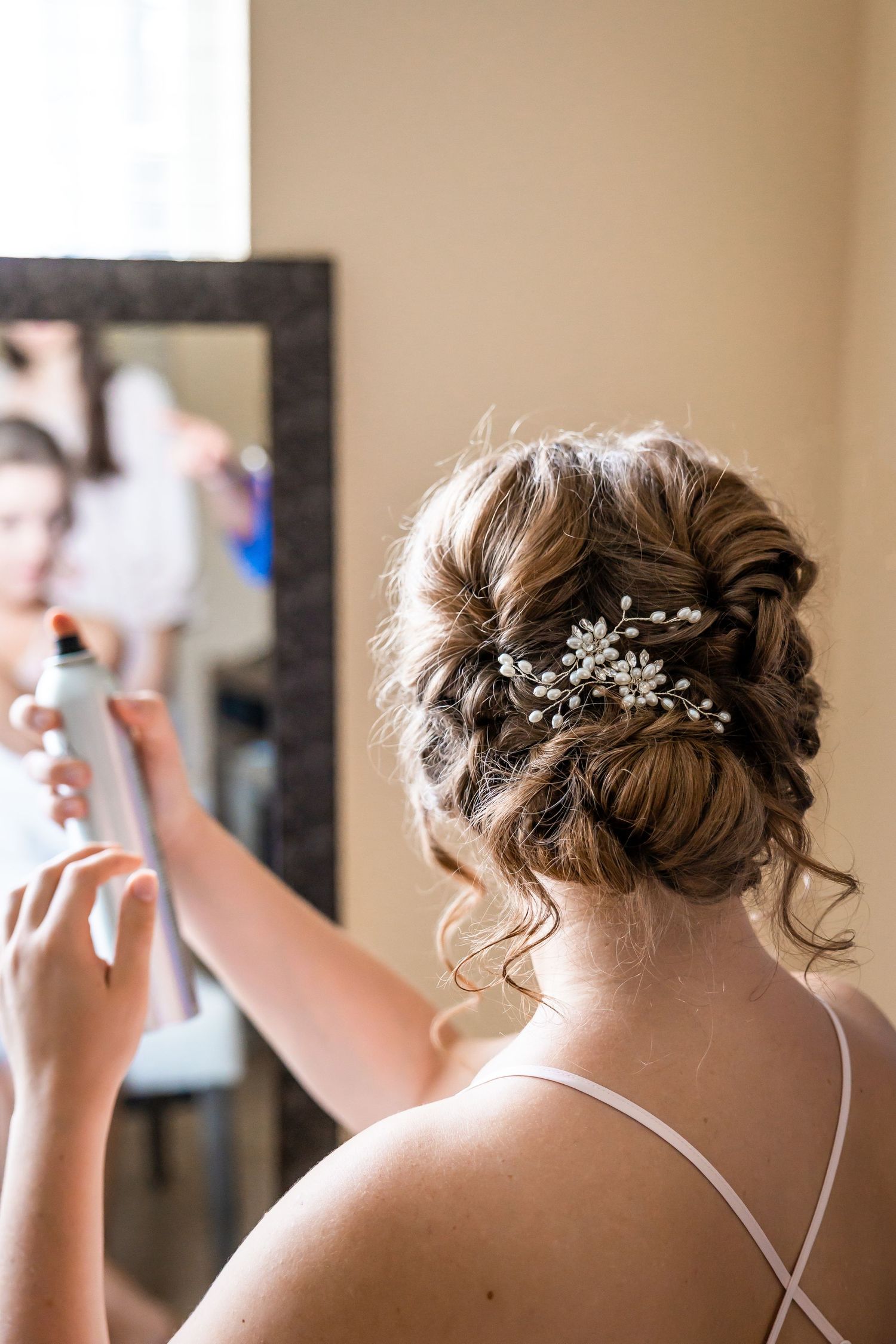 Stylist sprays hairspray on bride's elegant updo adorned with a pearl and crystal hair accessory in front of a mirror.