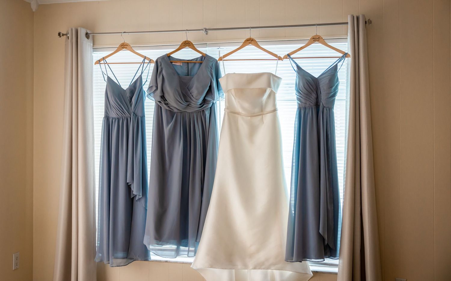 White wedding gown and three blue-gray bridesmaid dresses hang on wooden hangers in front of a bright window.