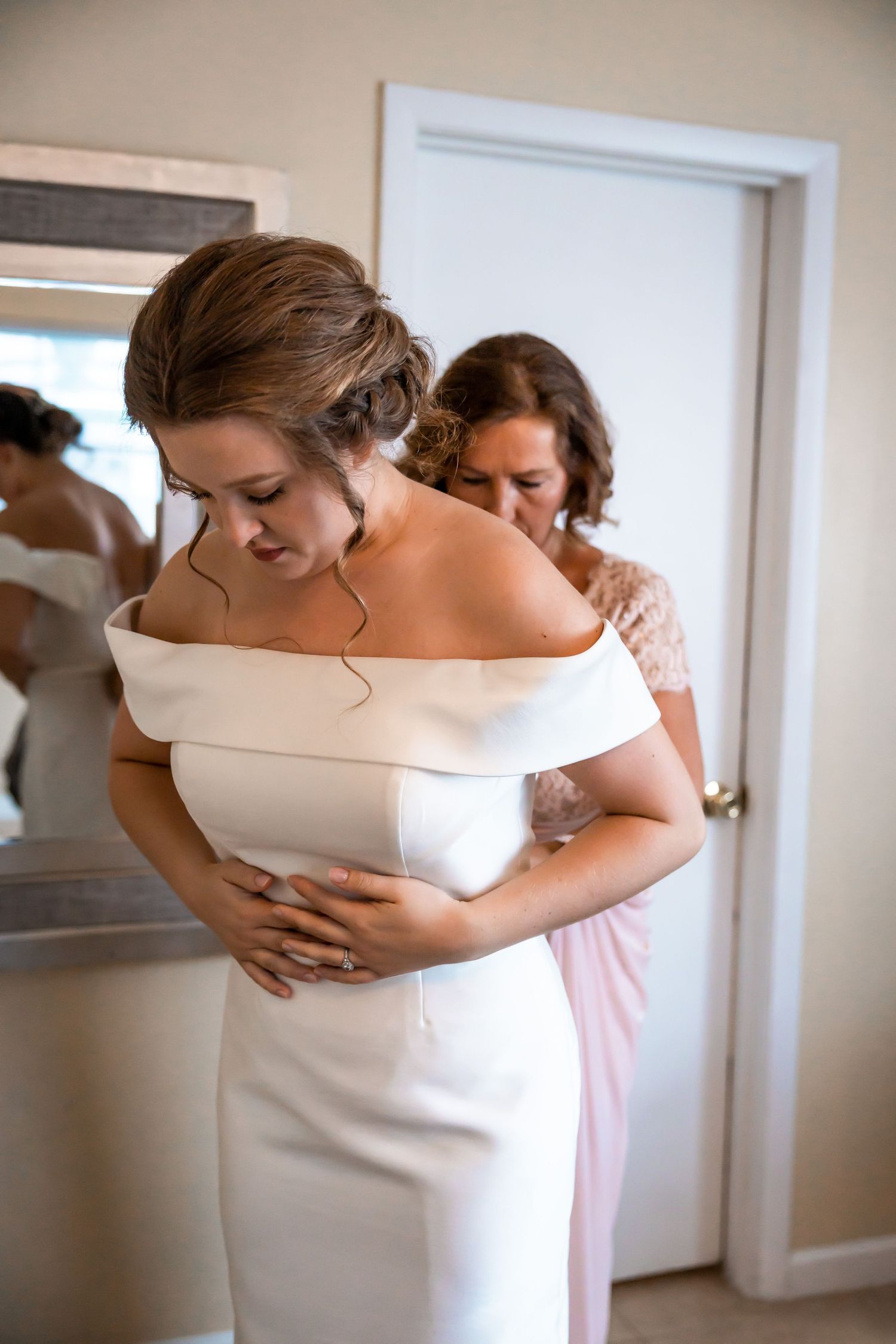 Bridesmaid helping bride zip up elegant off-shoulder white wedding gown before ceremony.