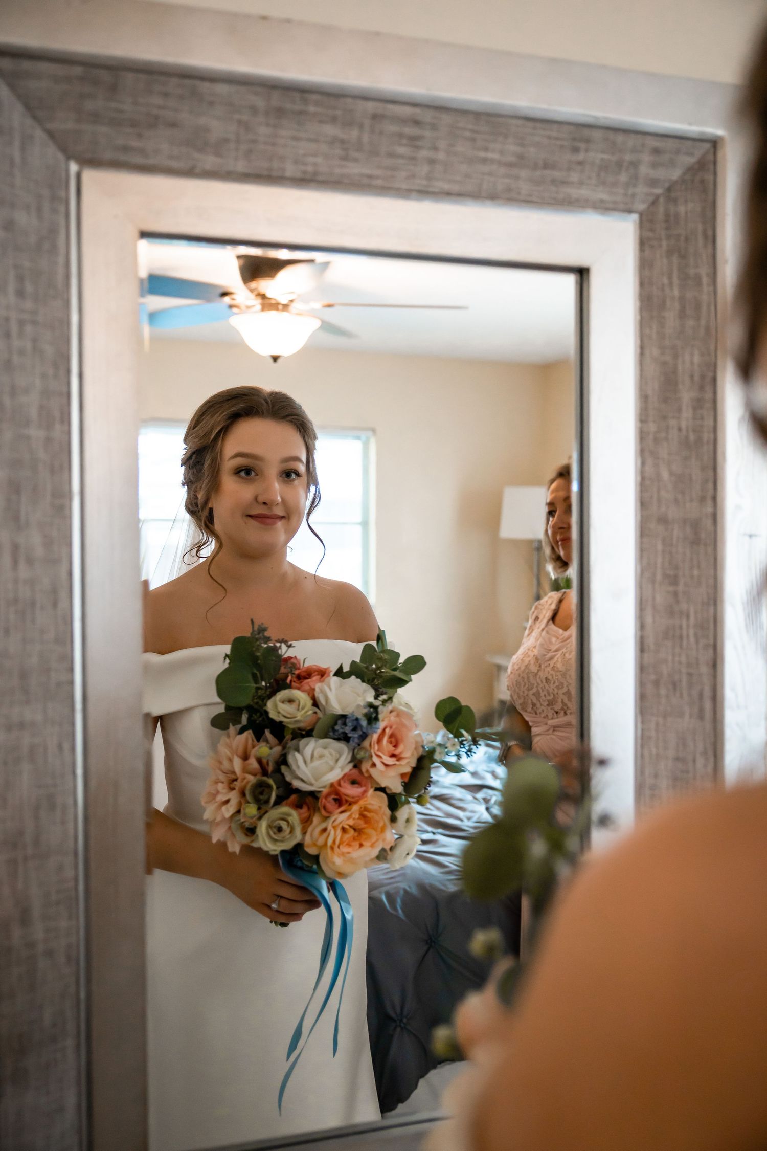 Bride in off-shoulder gown holding colorful bouquet reflected in large mirror.