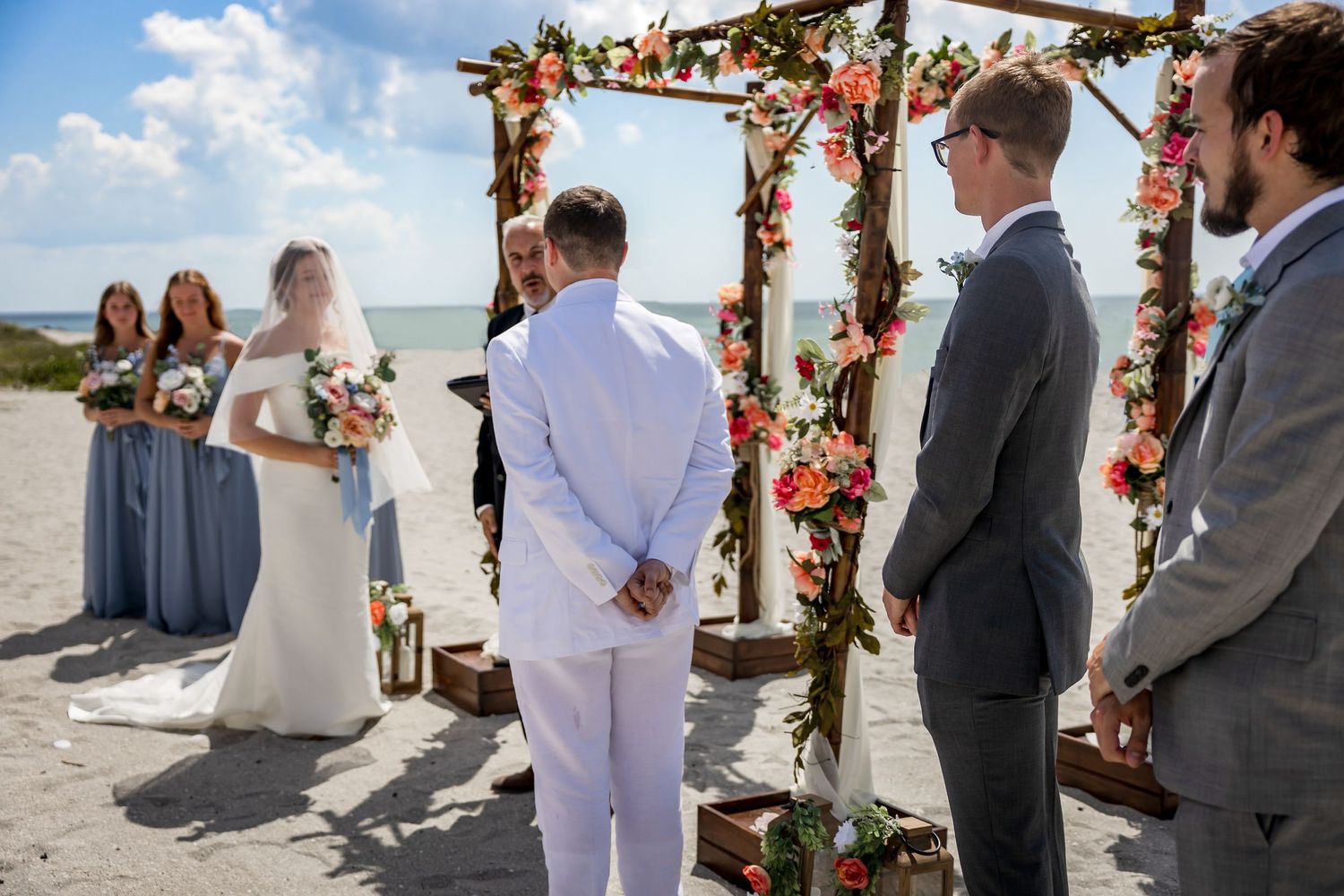 Beach wedding ceremony with floral arch, bridesmaids, groomsmen, and officiant by the ocean.