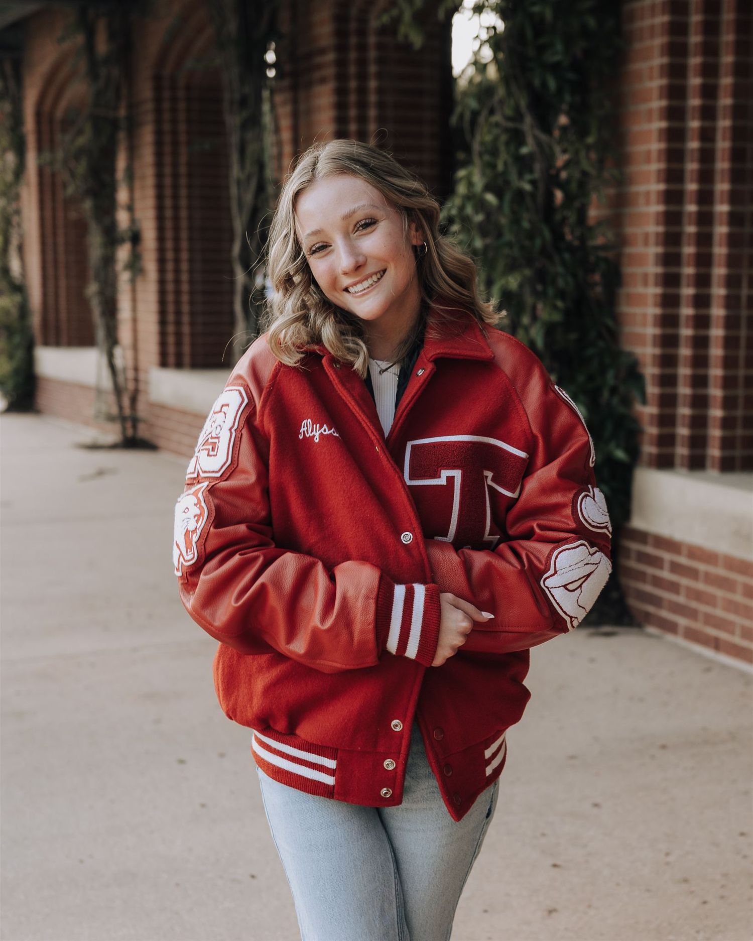 Casual pose in a red letterman jacket and jeans against classic brick architecture.
