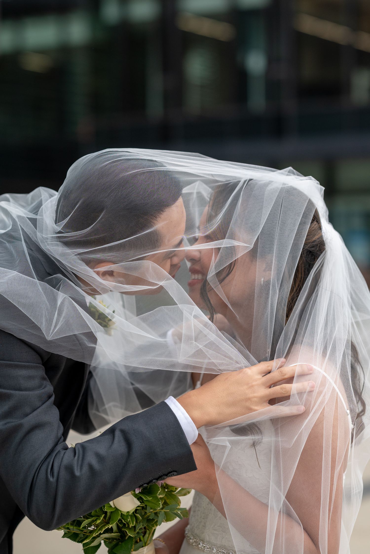 Romantic wedding moment captured under a flowing white veil against a modern building backdrop.