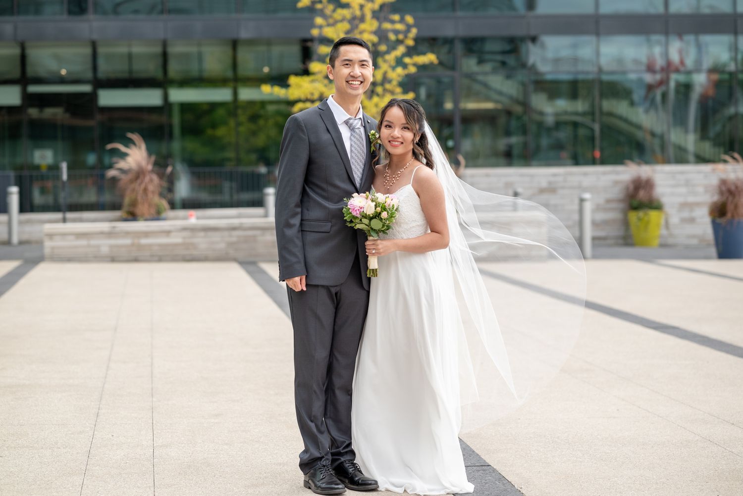 Elegant wedding couple poses outdoors in front of modern building architecture.