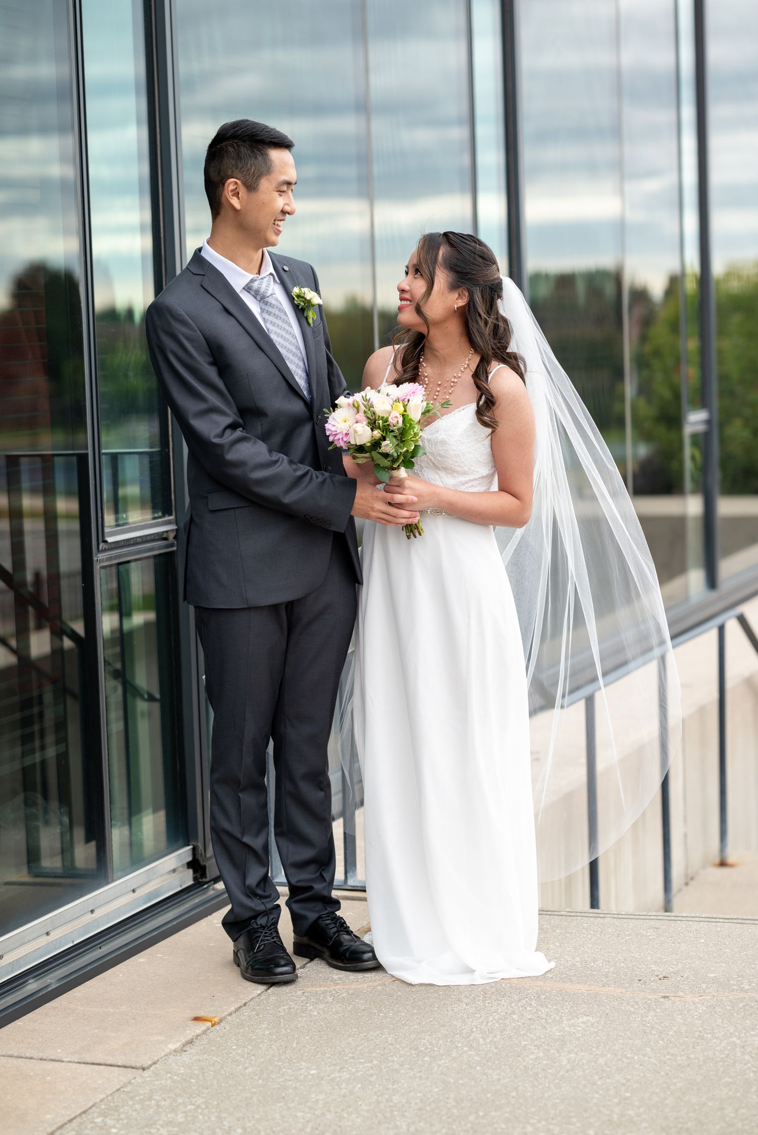 Wedding couple poses with bouquet against glass building exterior on their special day.