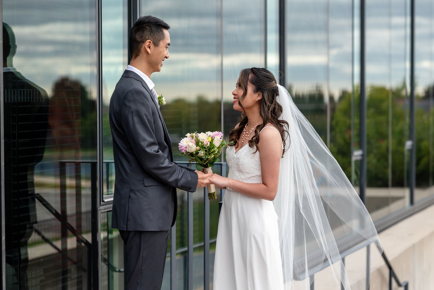 Wedding couple shares intimate moment with bouquet in front of glass building.