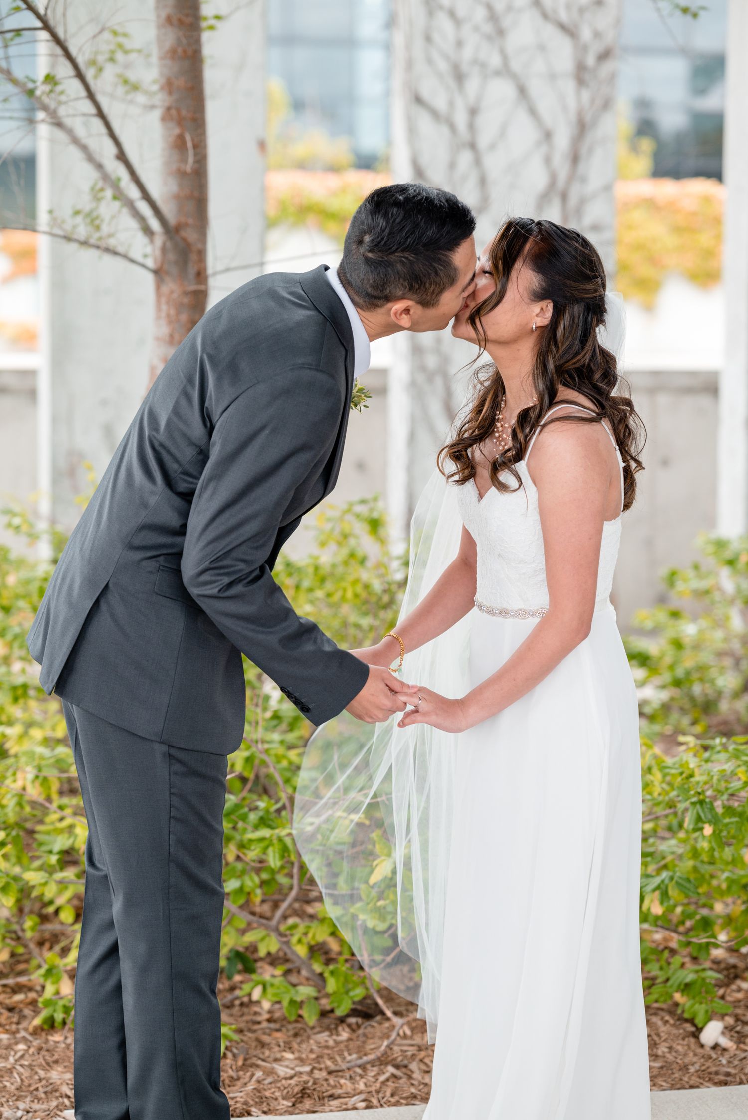 A romantic wedding moment captured among trees and landscaping in an outdoor garden setting.