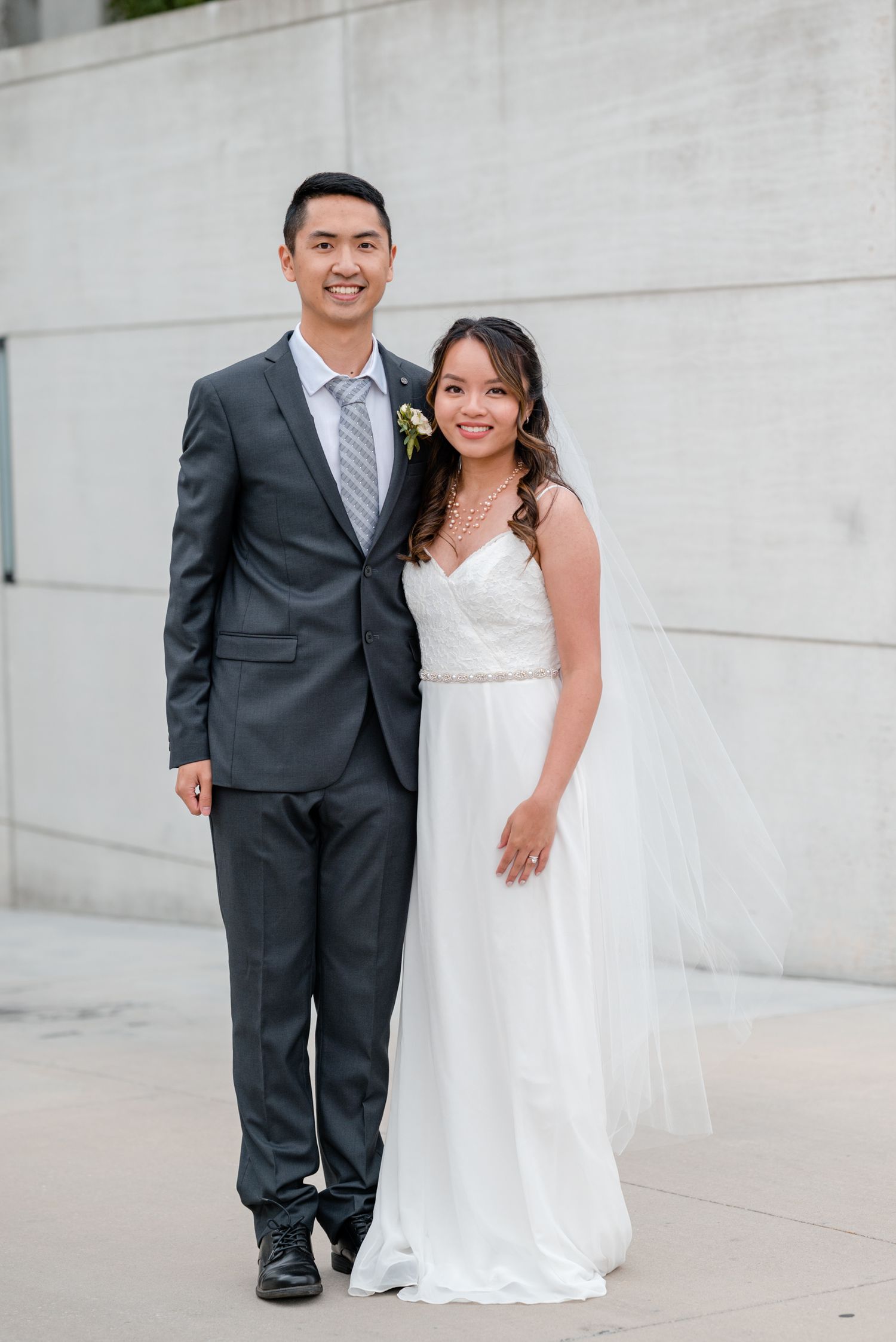 A bride in a white wedding gown and groom in a gray suit pose for a formal portrait against a concrete wall.