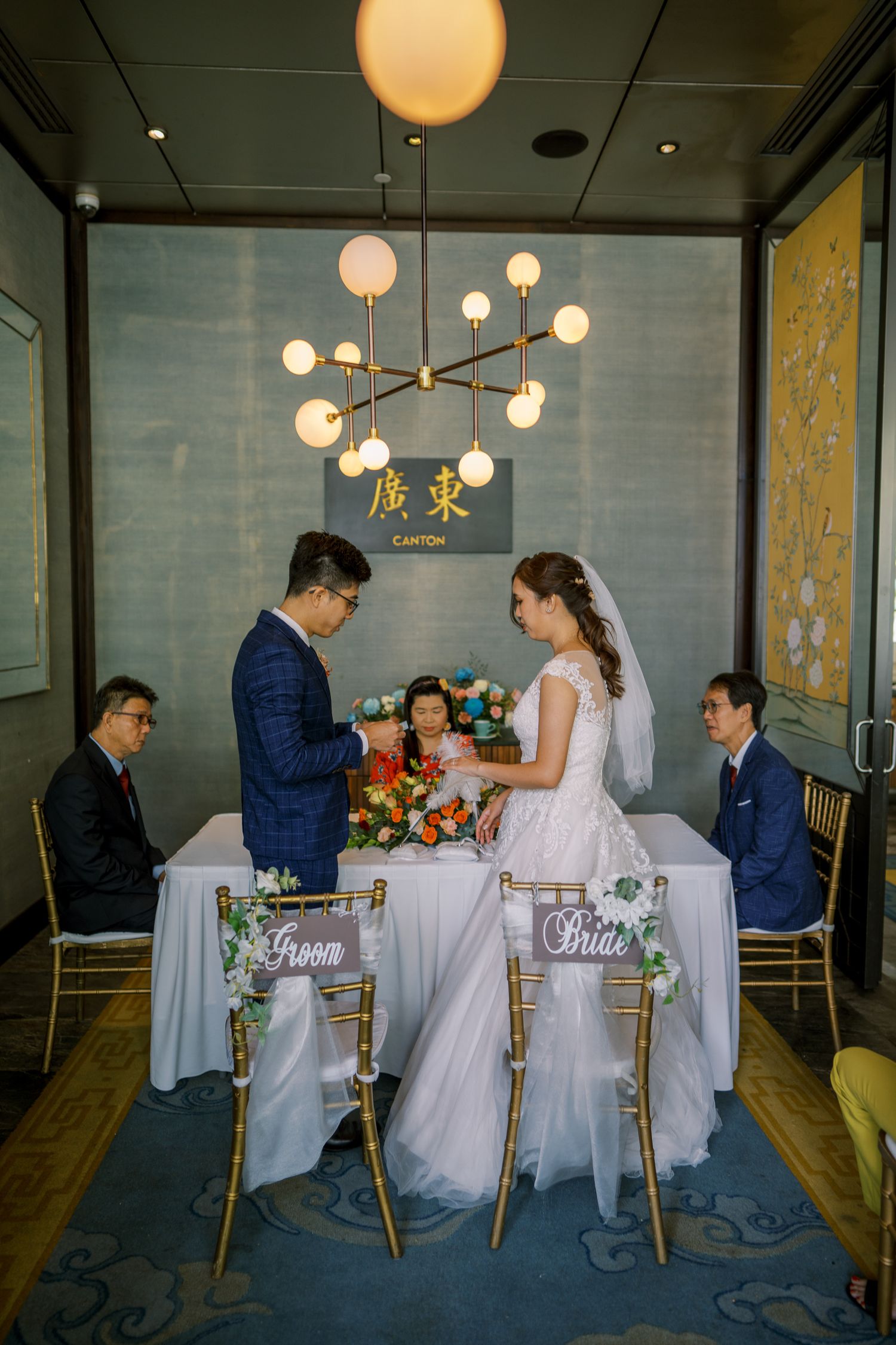 A wedding ceremony under modern globe pendant lights with bride and groom chairs labeled.
