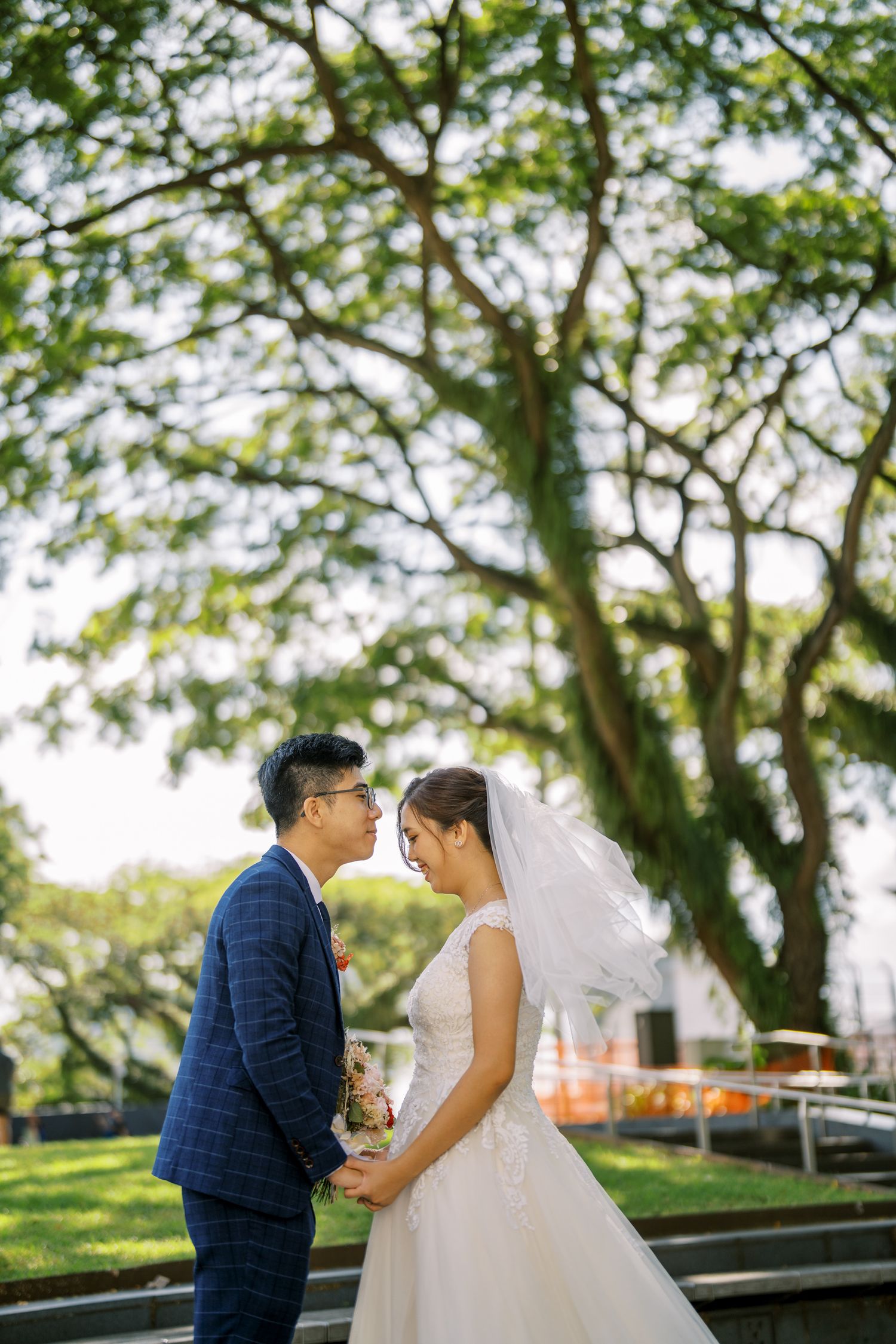 A romantic wedding couple shares tender moments in a lush garden setting with green trees in the background.