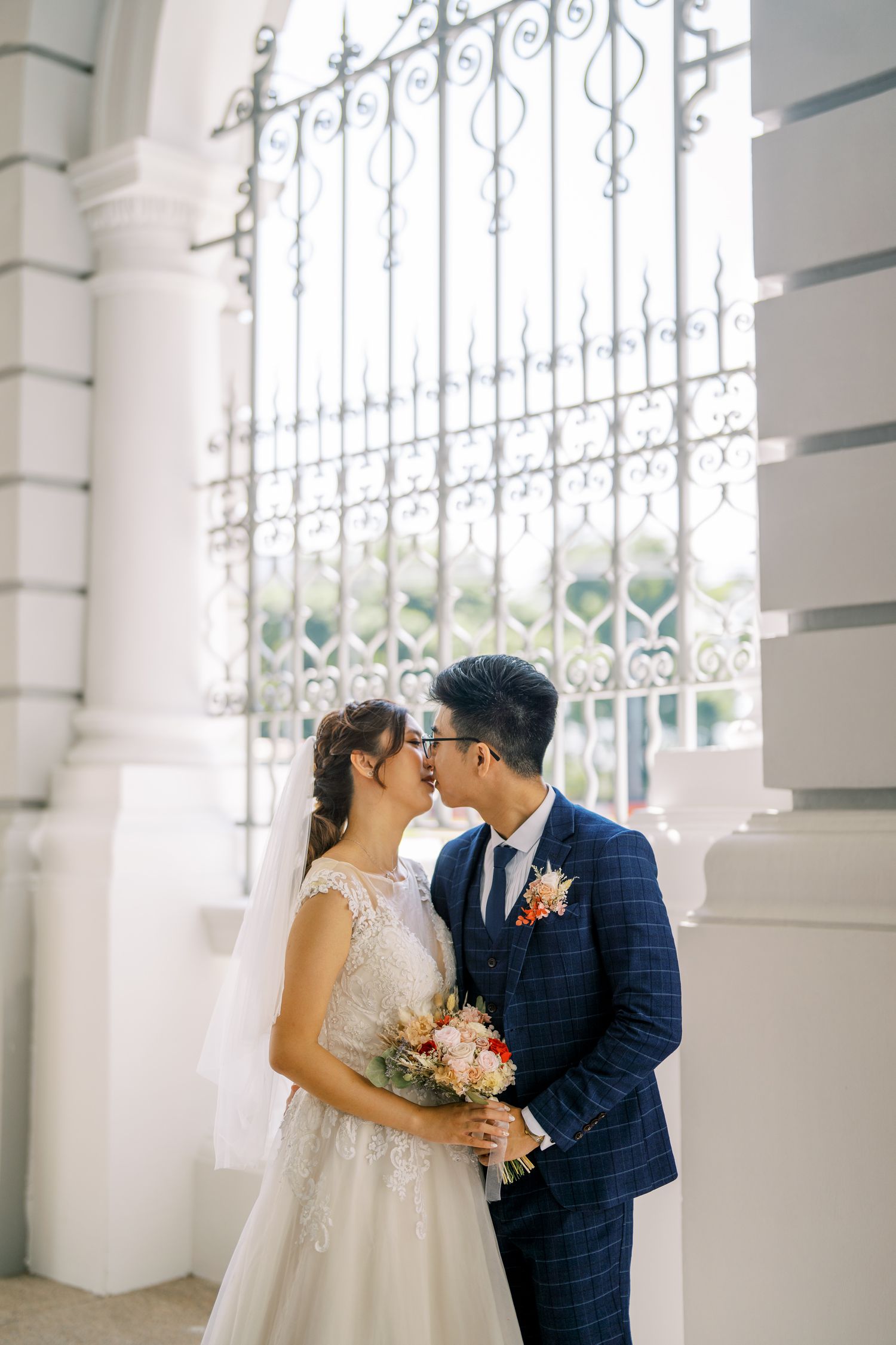A wedding couple embraces under elegant white columns and ornate iron gates.