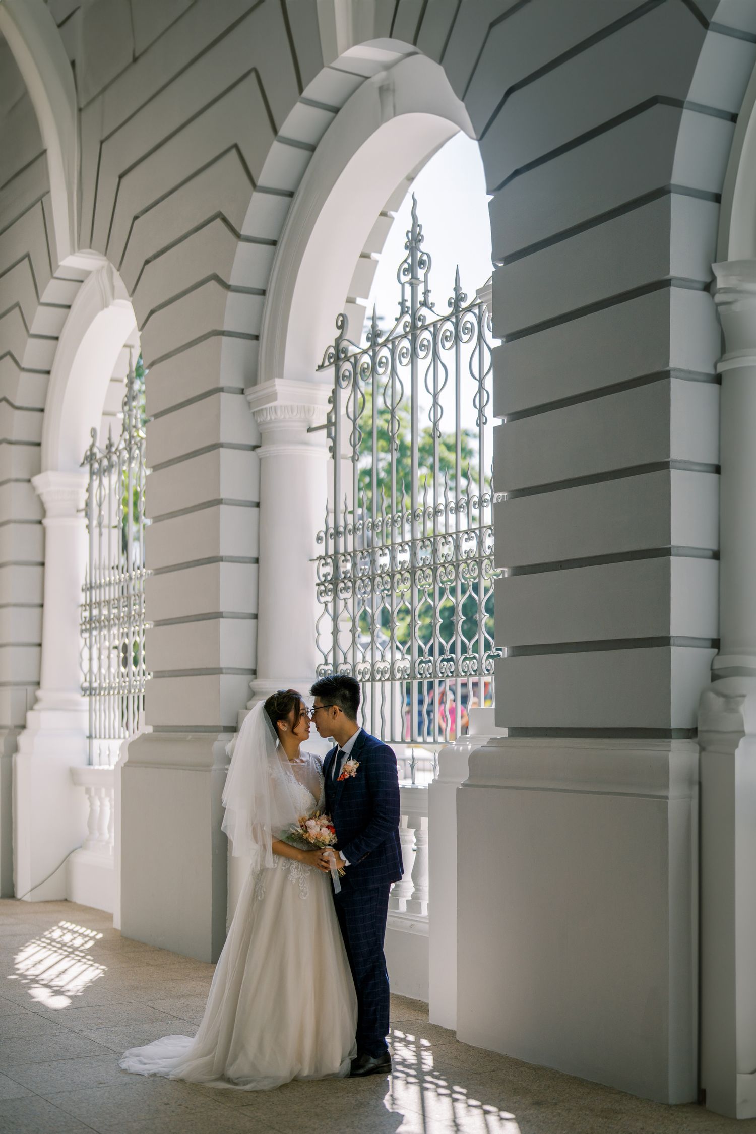 Newlyweds share intimate moments in a classic colonnade with arched windows and iron details.