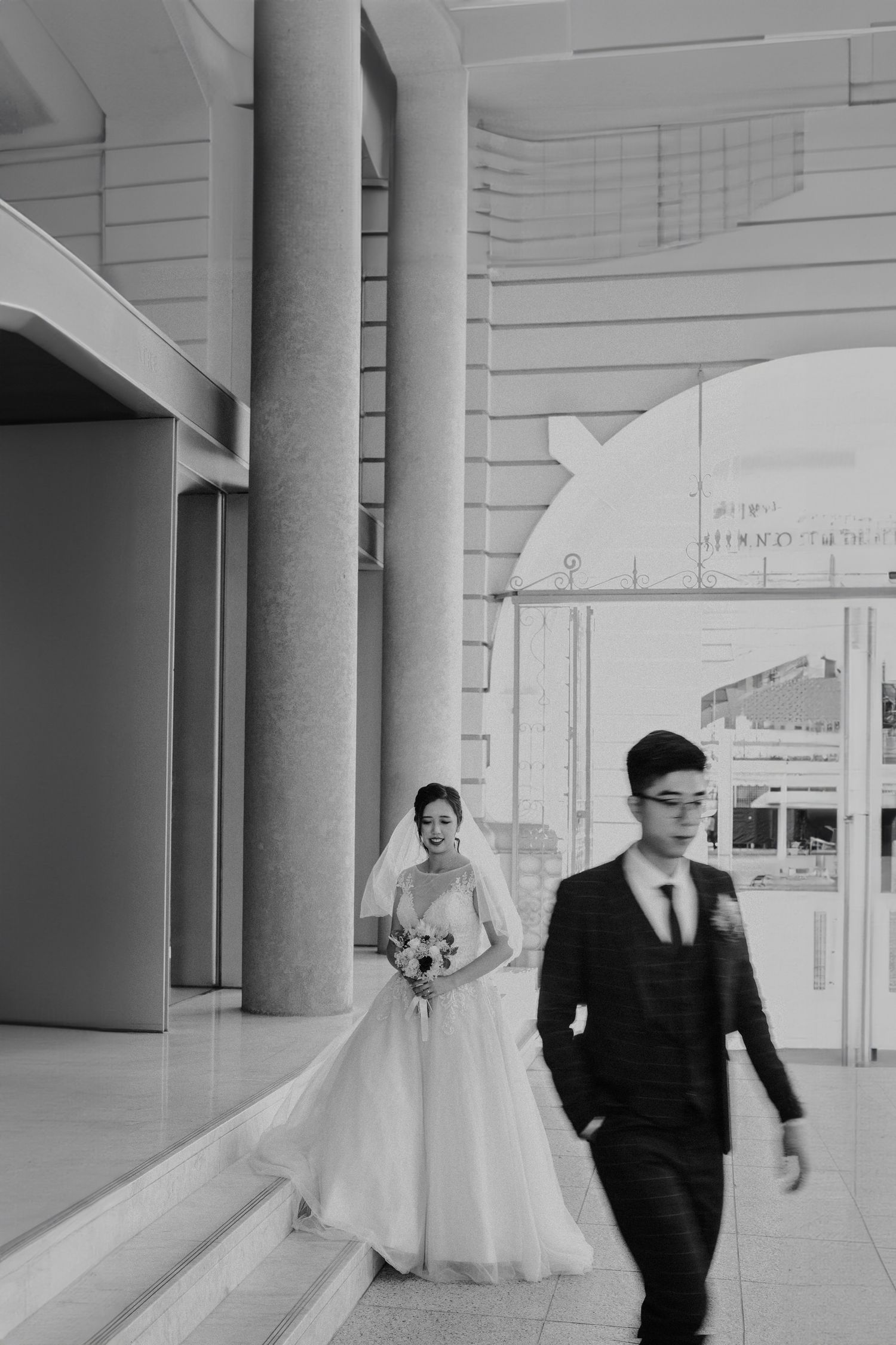 A wedding couple walks through a bright modern building with white columns.