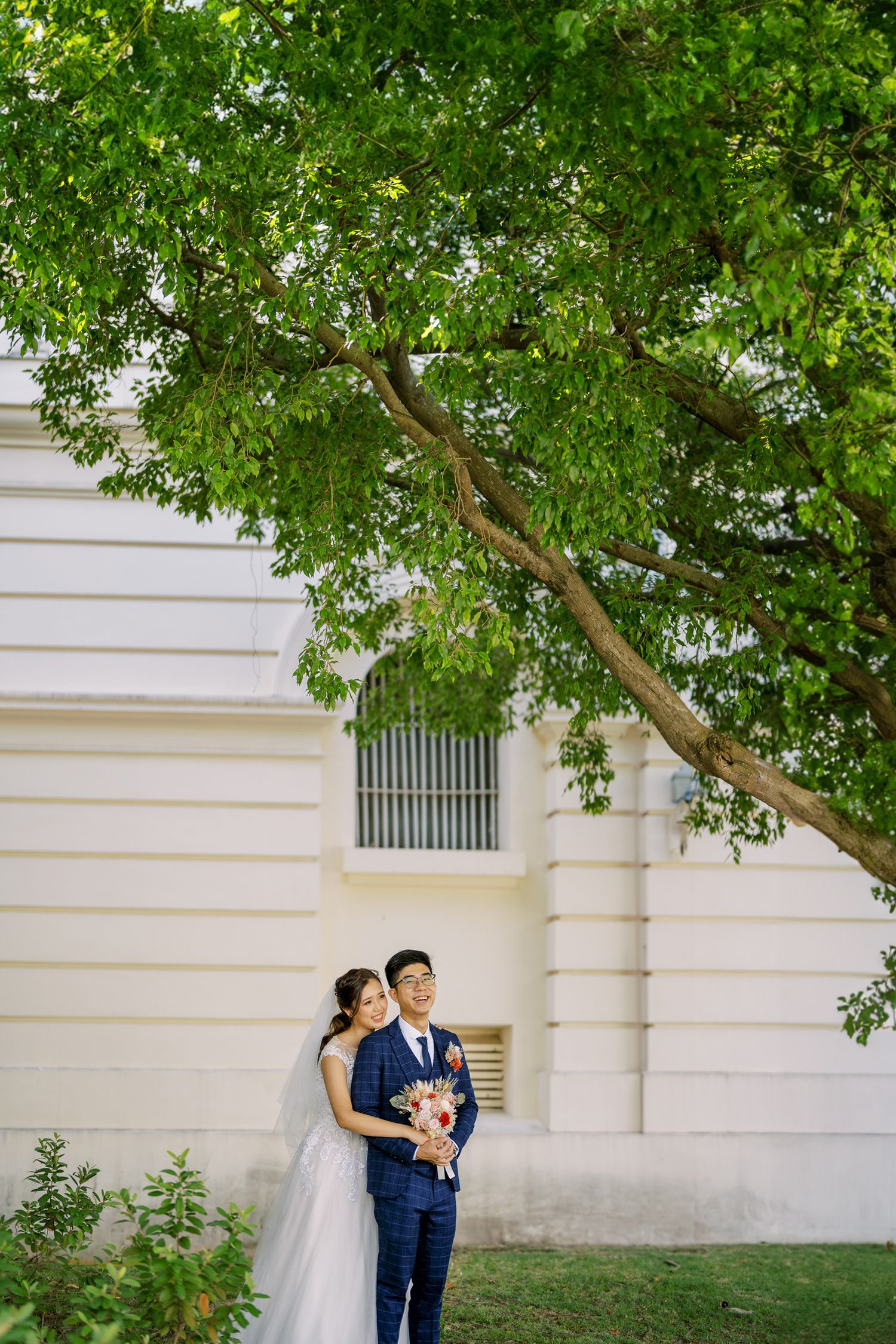 A couple poses for wedding photos against a white building with overhanging green trees.