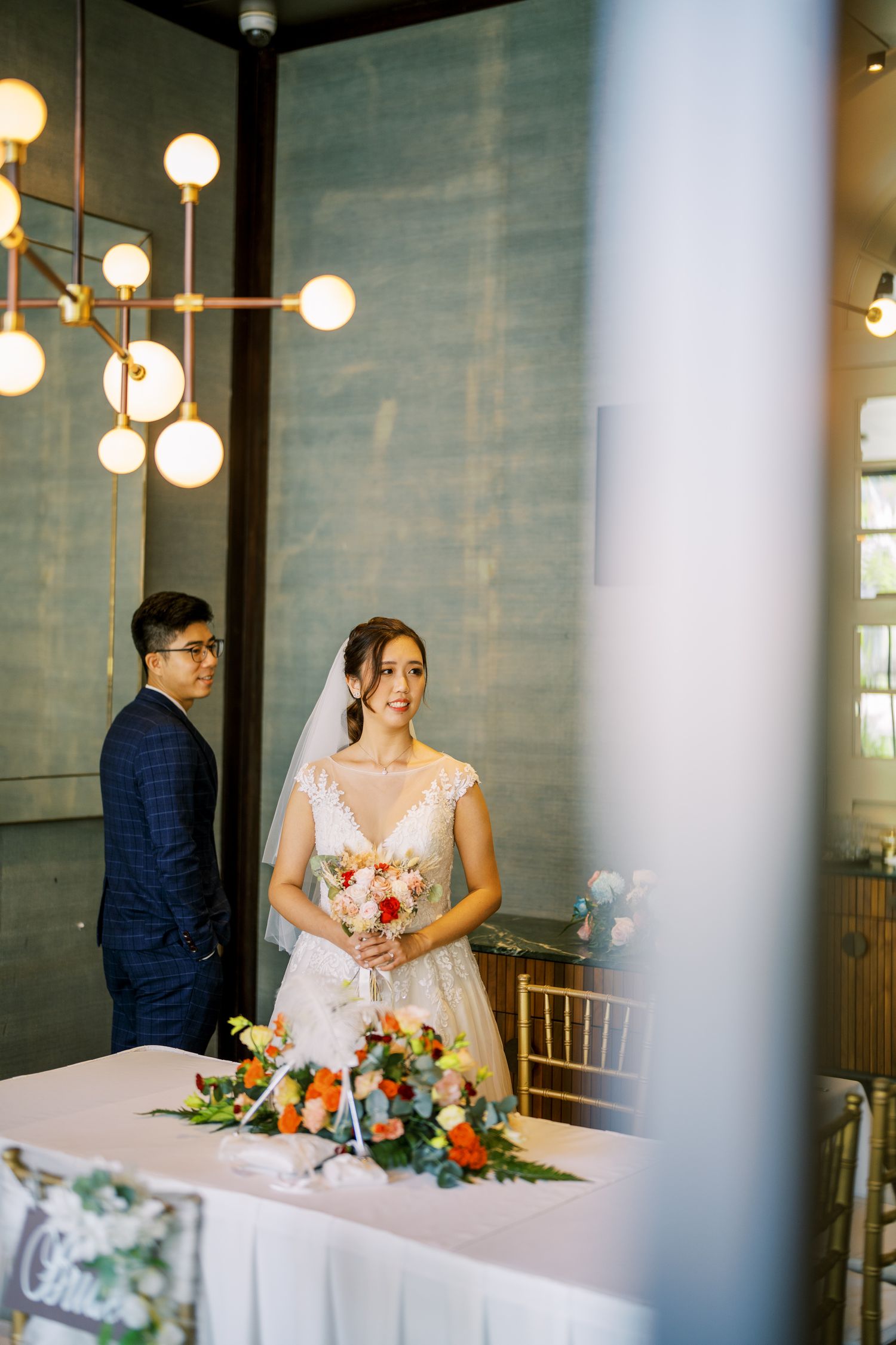 Wedding reception scene through columns with modern lighting and elegant table settings.