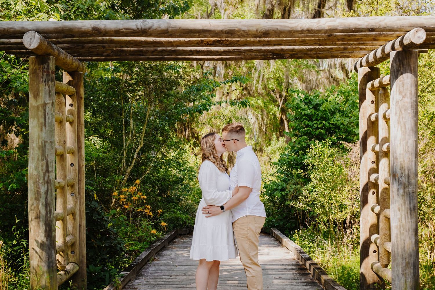 A romantic couple embraces on a rustic wooden bridge surrounded by lush greenery at a park.