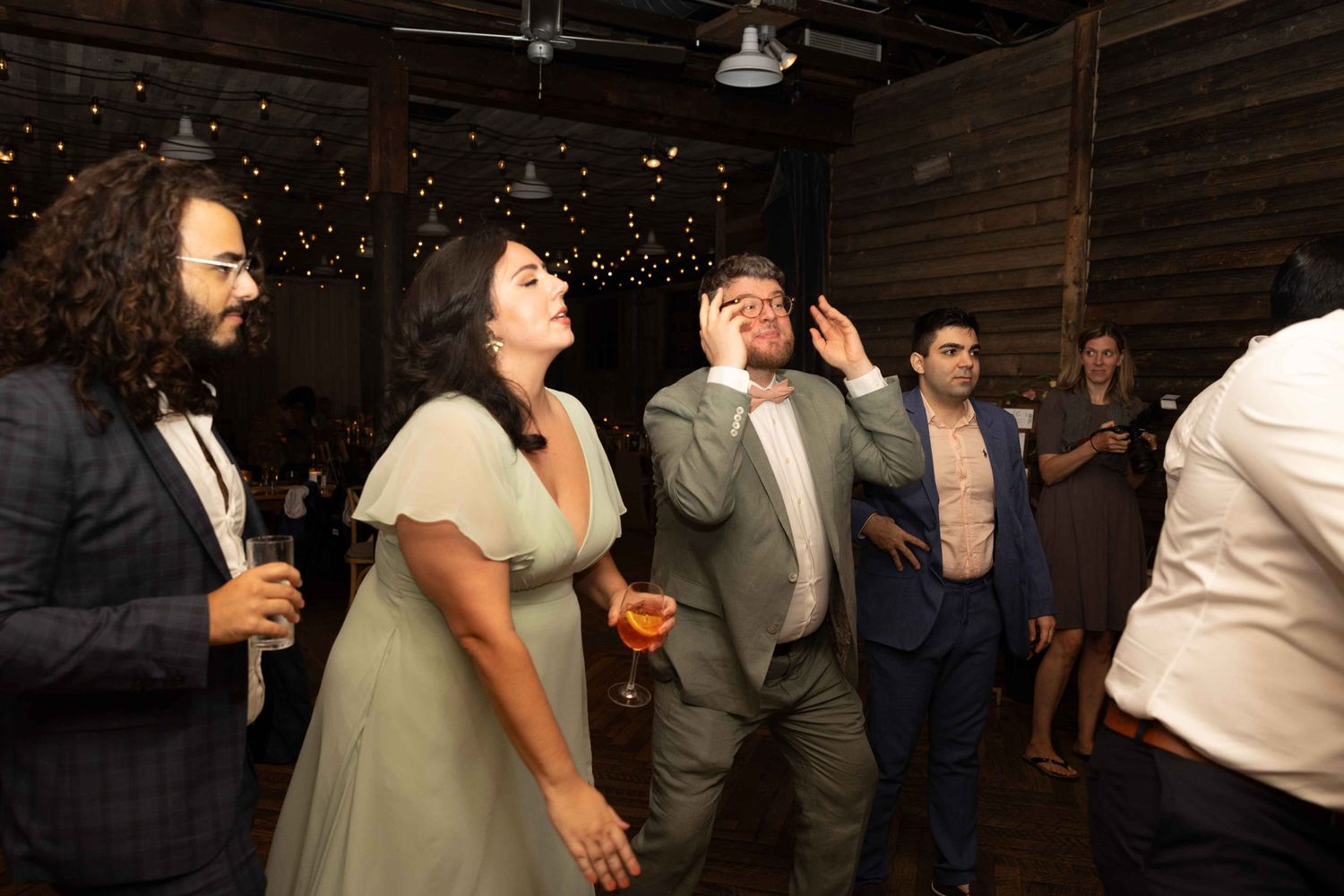 Party guests looking upward at decorative lights in a rustic venue with wooden beams.