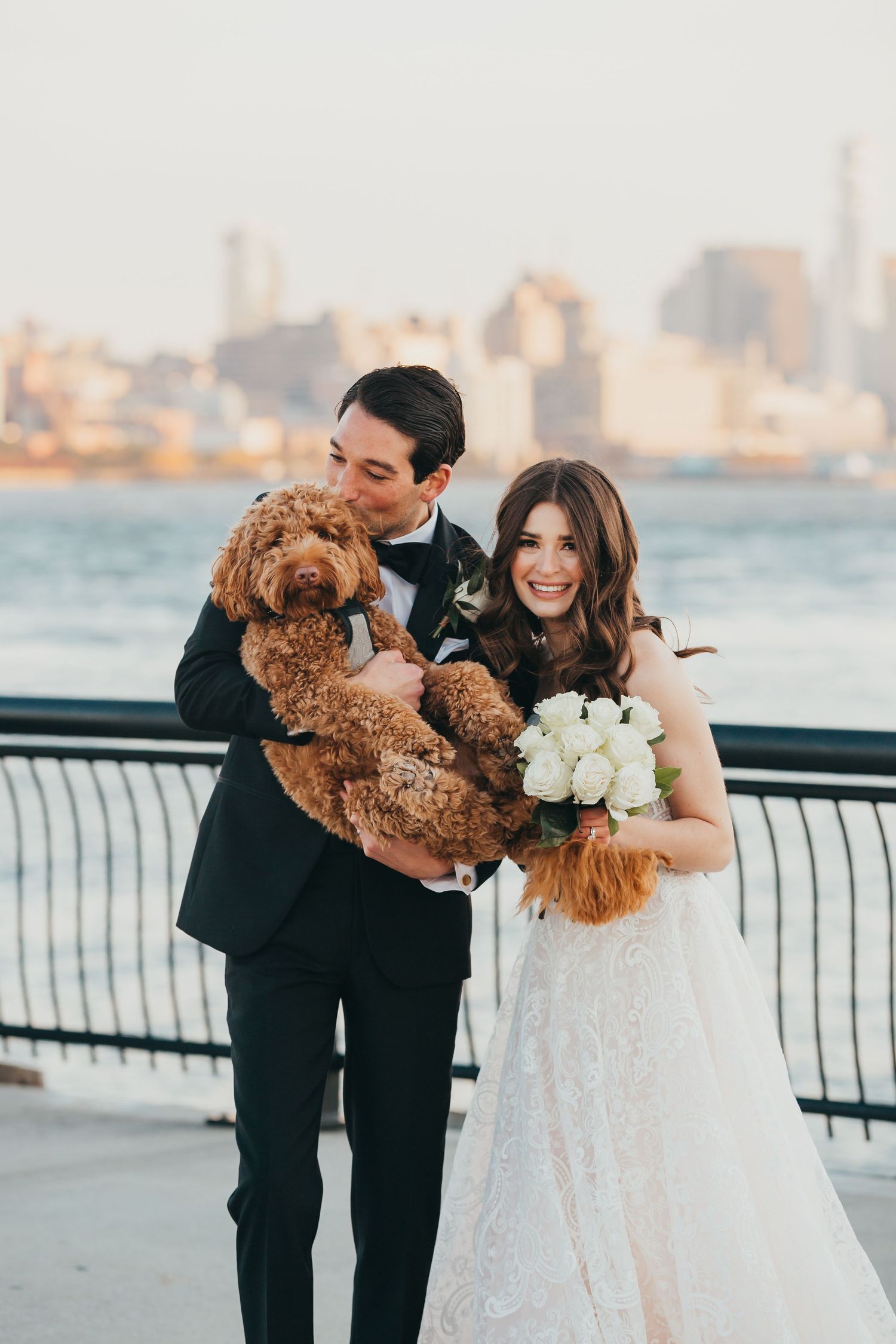 Smiling bride and groom holding an adorable brown doodle dog with white roses and city skyline.