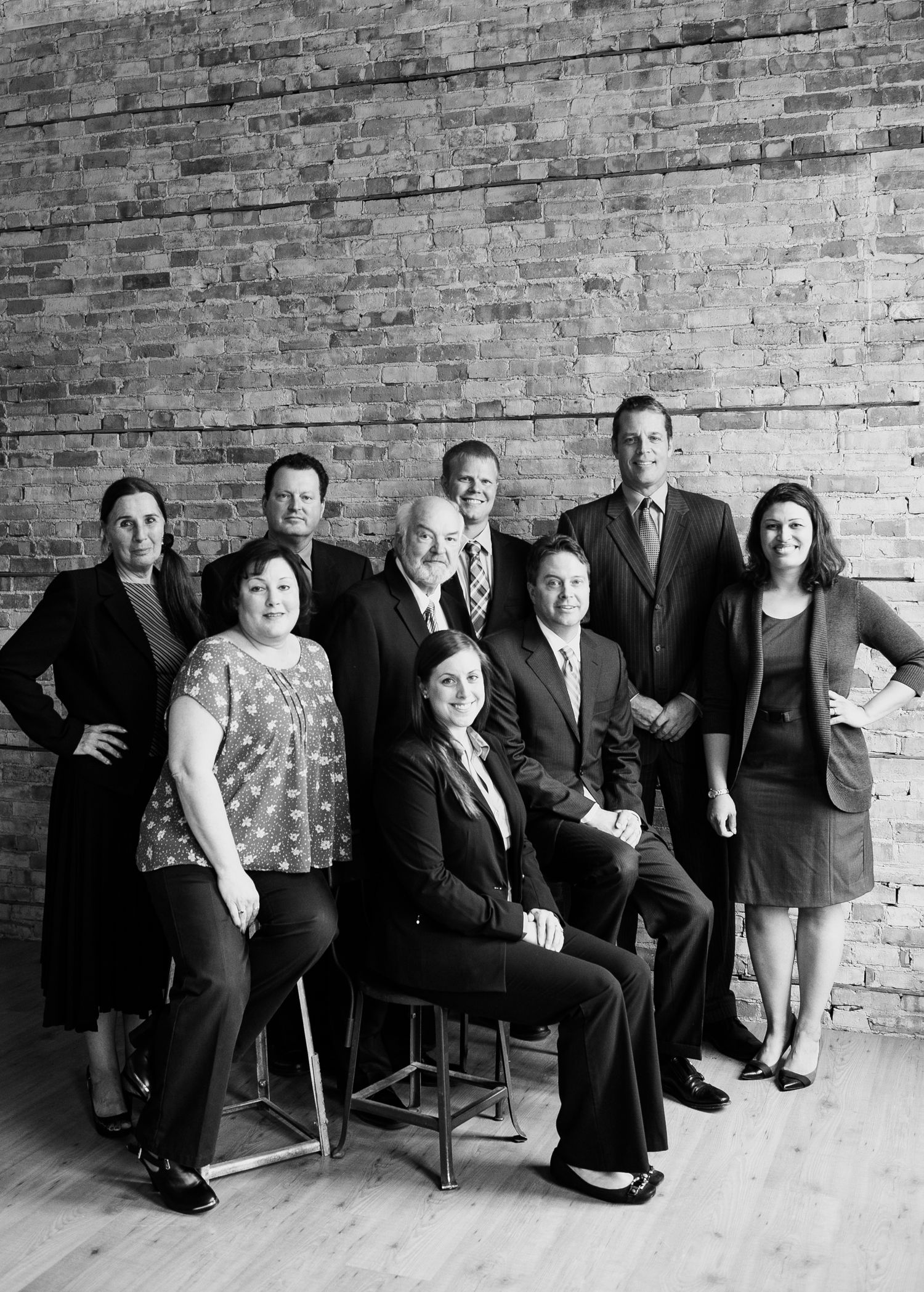 Black and white professional group portrait of nine business professionals posed against brick wall.