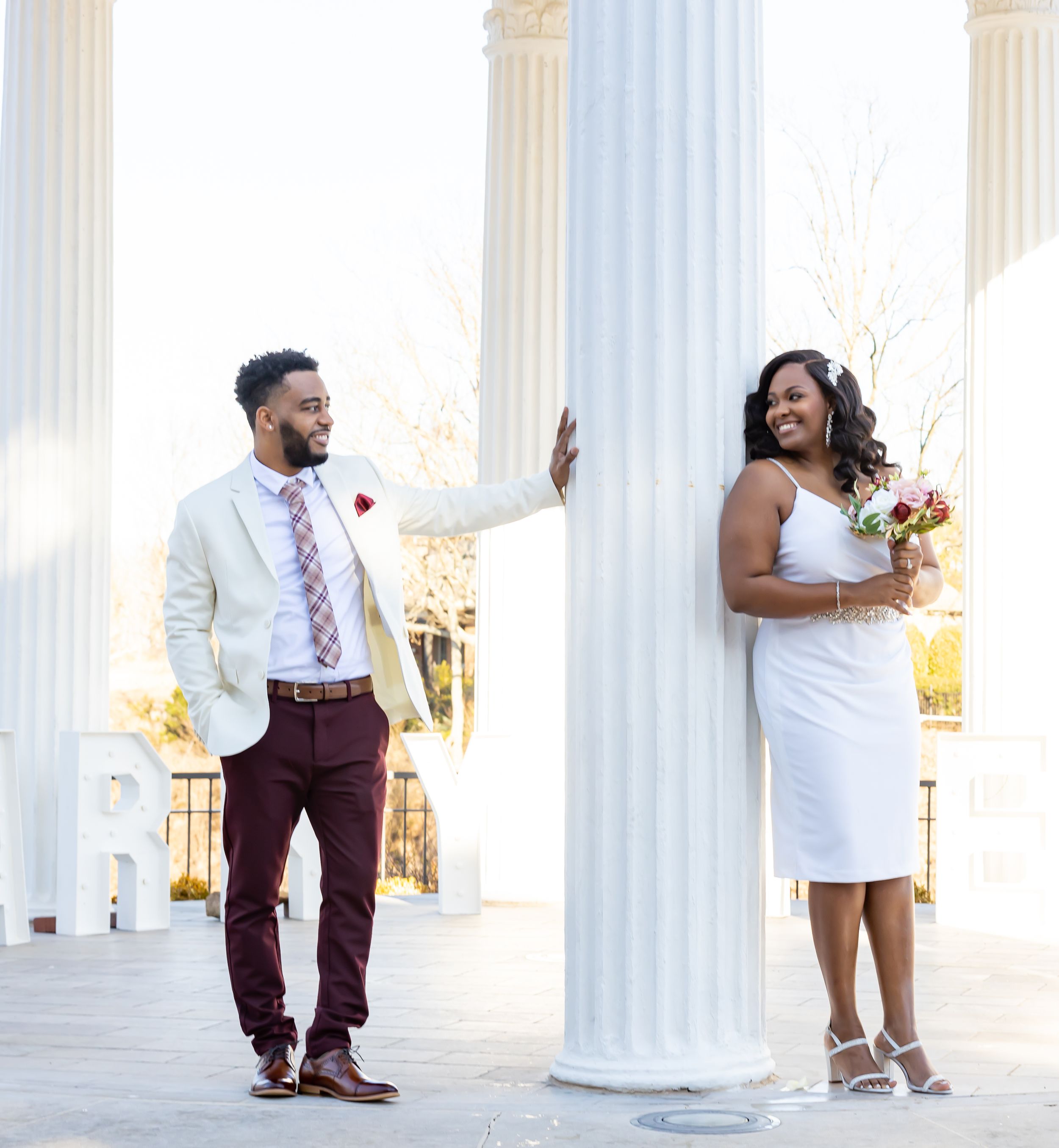 Couple poses playfully between white columns in formal wedding attire.