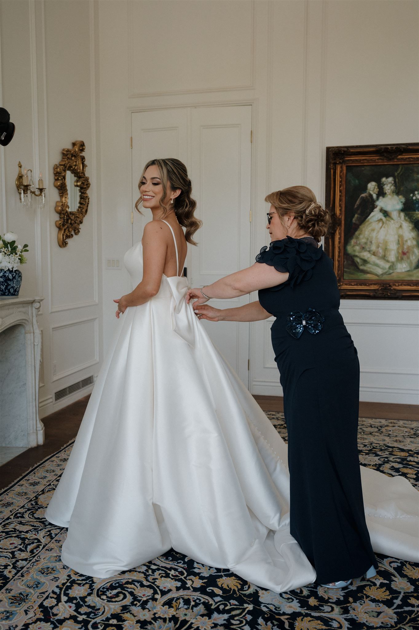 Final preparations of a beautiful white wedding dress being adjusted in an elegant room with ornate decor and artwork.