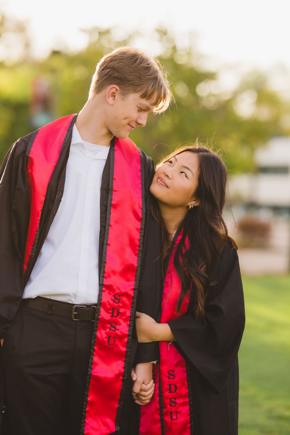 SDSU couple walking hand in hand across the campus lawn in graduation outfits.