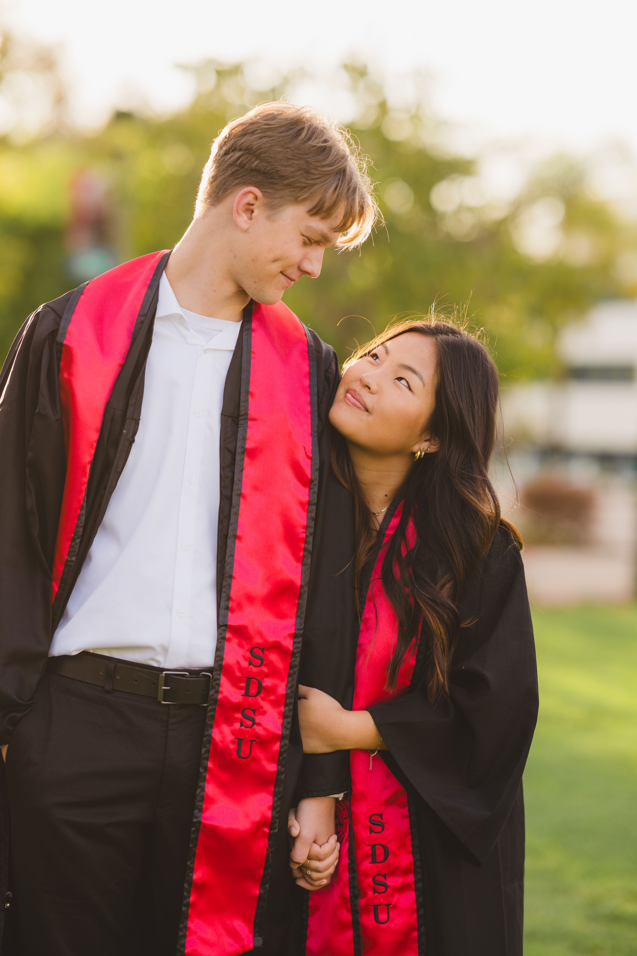 SDSU couple walking hand in hand across the campus lawn in graduation outfits.