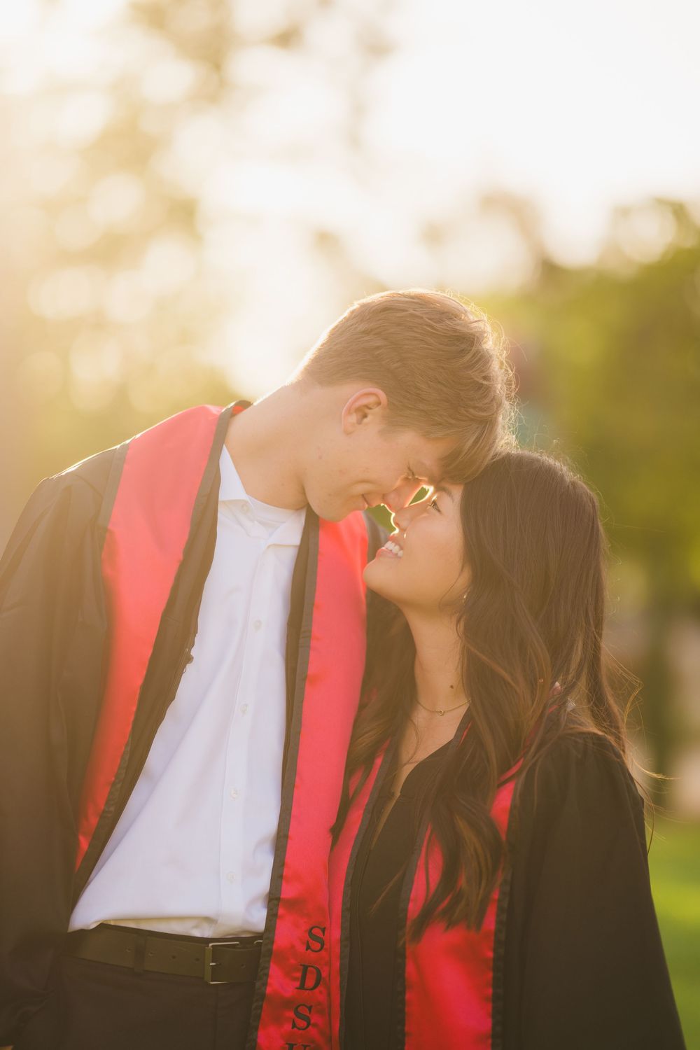 SDSU graduating couple in cap and gown touching foreheads and smiling softly in front of Hardy Tower.
