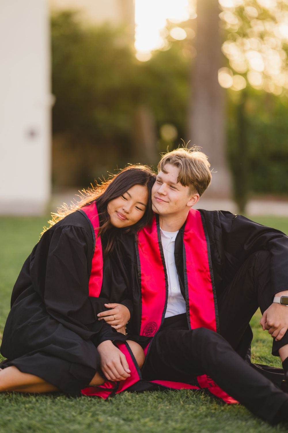 SDSU senior holding hands with partner while both smile at the camera in cap and gown while sitting on the grass.