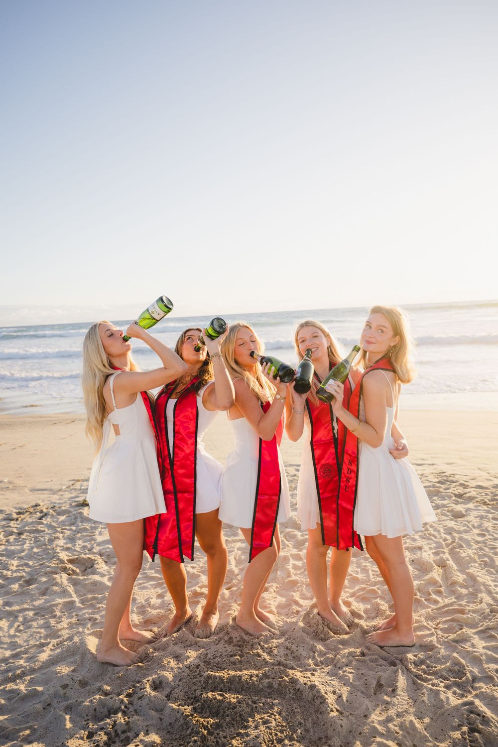 Group of SDSU graduates wearing their stoles laughing and playing around barefoot in the soft sand at Mission Beach.