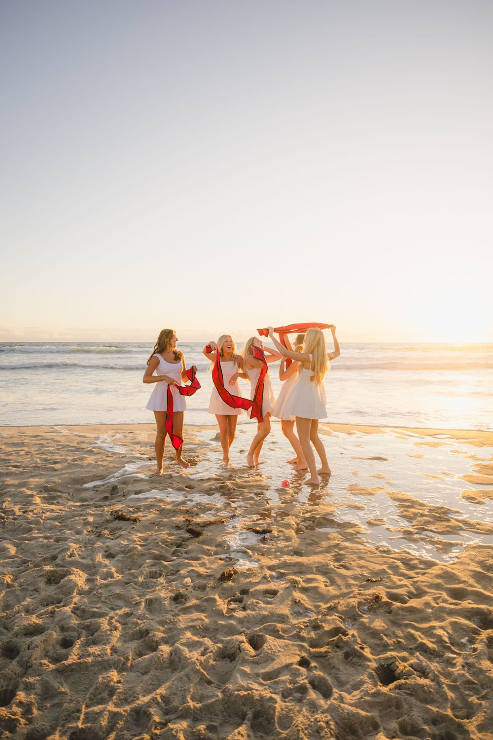 Group of SDSU graduates holding their stoles laughing and playing around barefoot in the soft sand at Mission Beach.