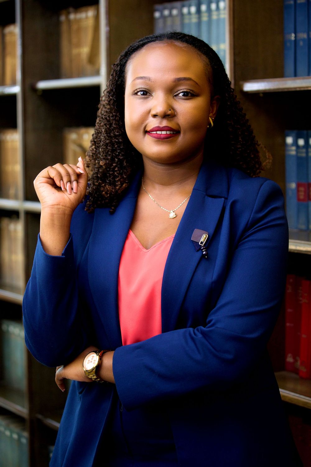 Professional portrait in navy blazer and coral top against library background.