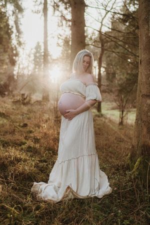 Maternity photo of woman in white dress standing in sunlit forest setting.