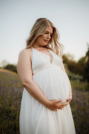 Side profile of pregnant woman in flowing white dress against light background.