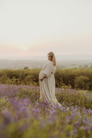 Pregnant woman in white dress stands among purple wildflowers during sunset.