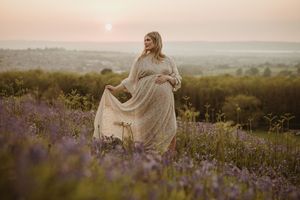 Maternity photo in flowing dress amid purple wildflowers during golden hour.