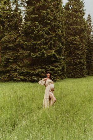 A maternity photo of a woman in a flowing dress standing in a grassy field near evergreen trees.