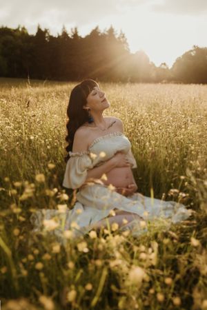 Expectant mother in white dress sitting among wildflowers during golden hour sunset.